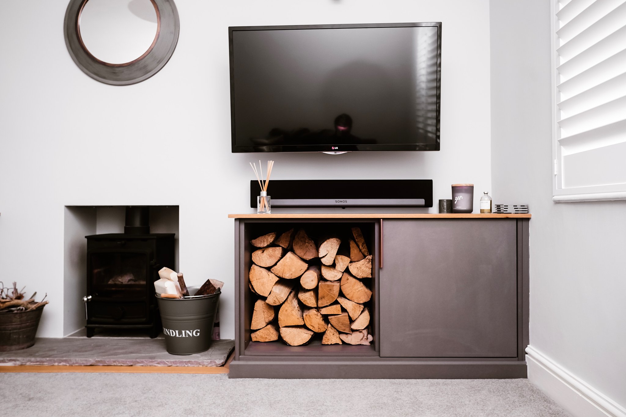 Living room with fireplace, wood logs in a storage compartment, TV mounted on wall, soundbar below TV, decoration items on cabinet, and window with white blinds.