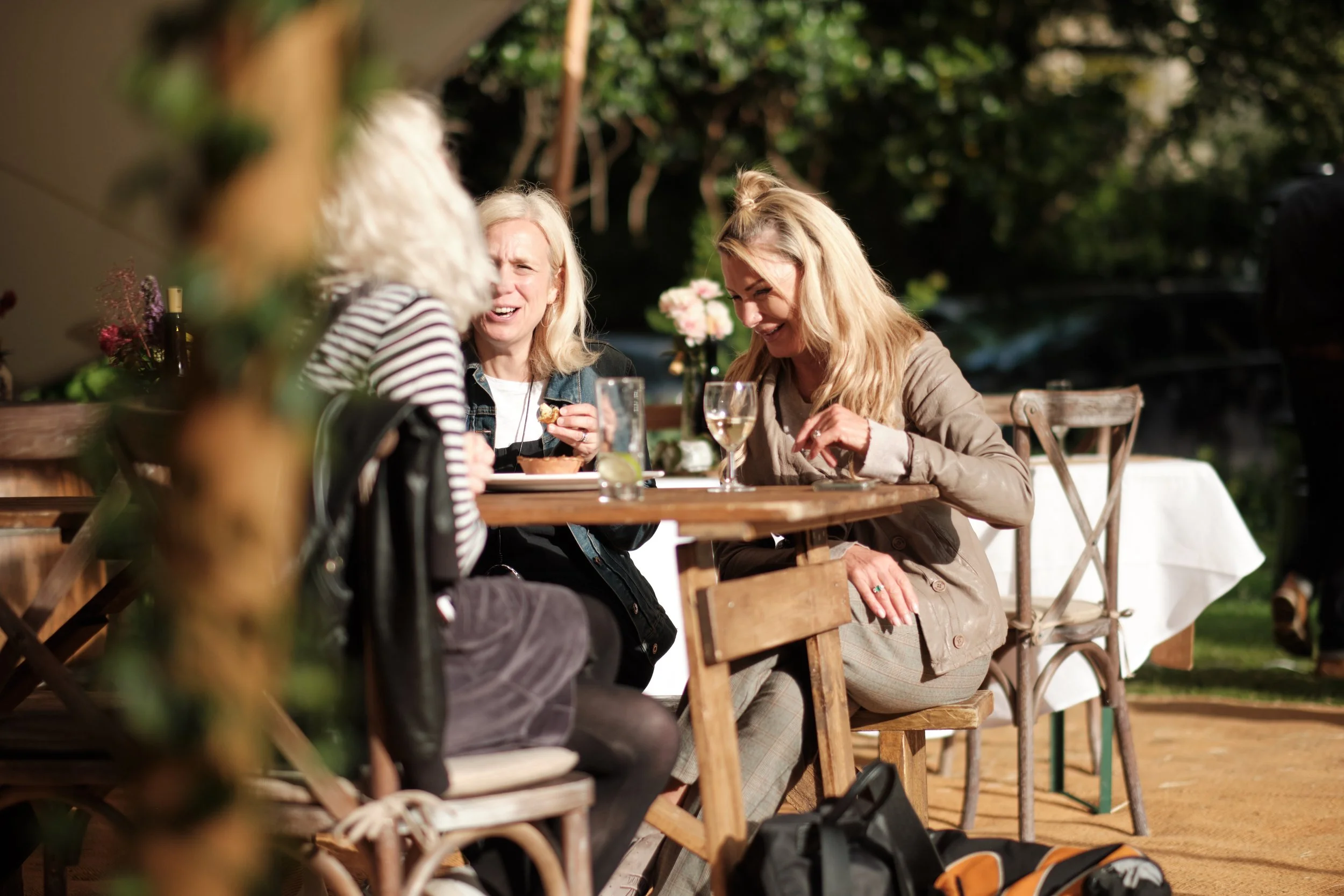 Three women are sitting at an outdoor wooden table, laughing and enjoying drinks and food on a sunny day.