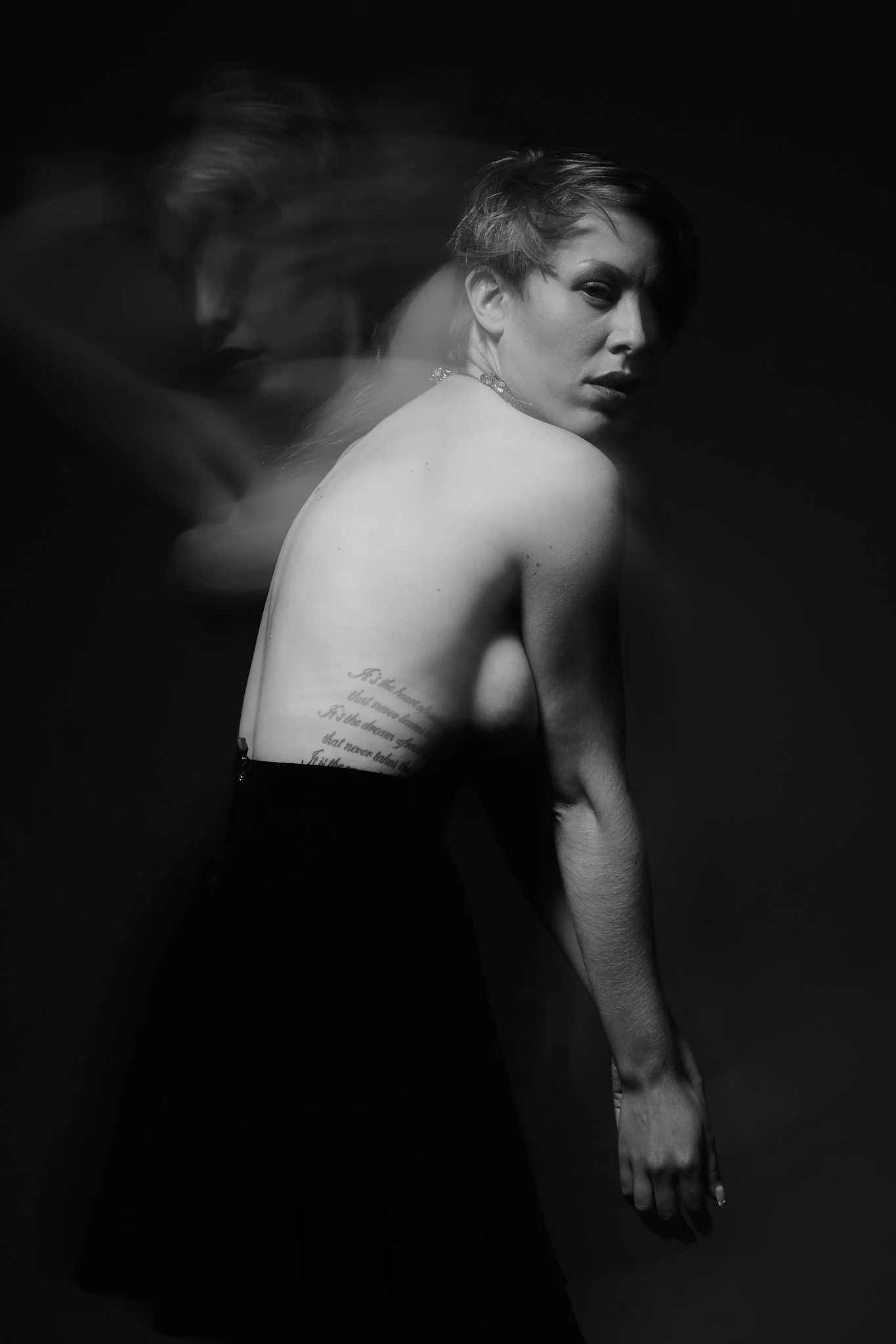Black and white portrait of a woman with short hair, facing sideways with her head turned toward the camera, partially covered in motion blur, with visible tattoo on her side, against a dark background.