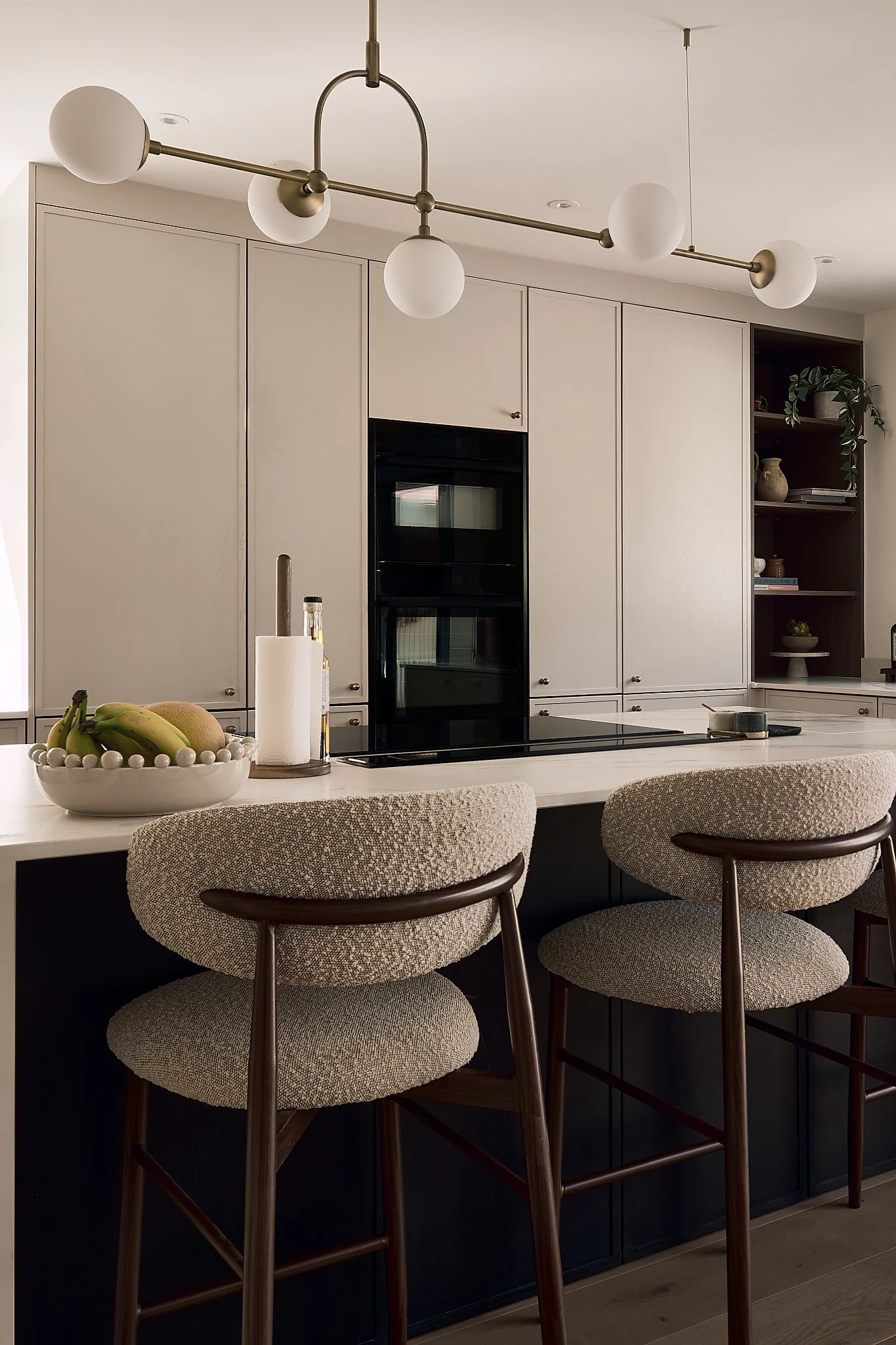 Modern kitchen with beige cabinets, black oven, and a white countertop. Three upholstered bar stools with wooden legs are placed in front of the counter. A bowl of fruit and paper towels are on the counter, and open shelves with decorative items are on the wall.