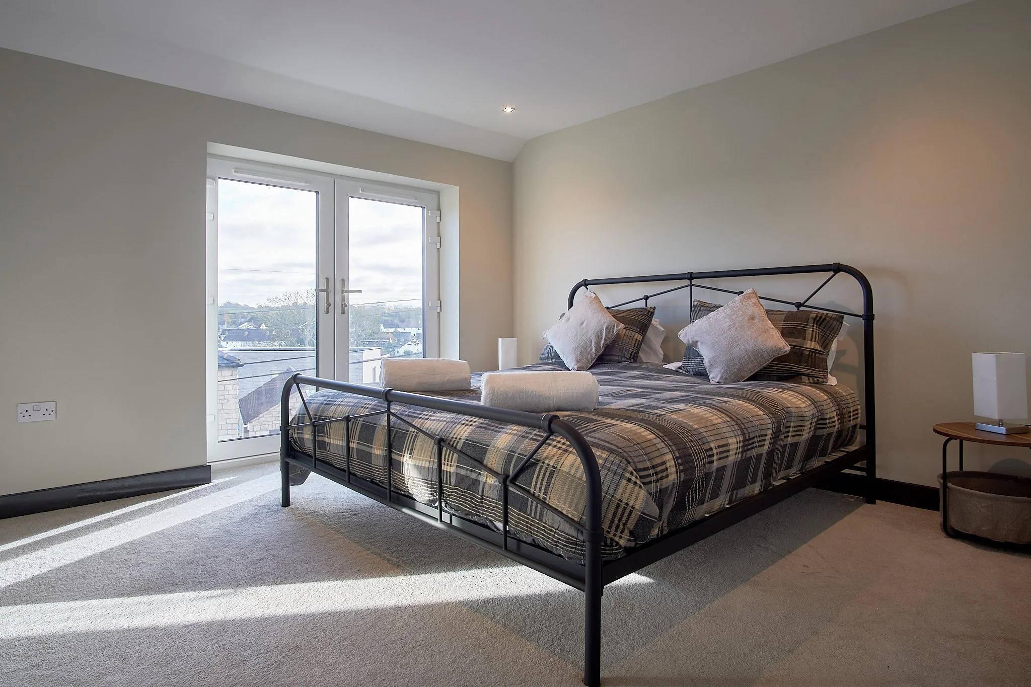 Bedroom with a black metal bed frame, plaid bedding, three pillows, two rolled towels, a nightstand with a lamp, and a large window showing a cloudy daytime sky and rooftops.