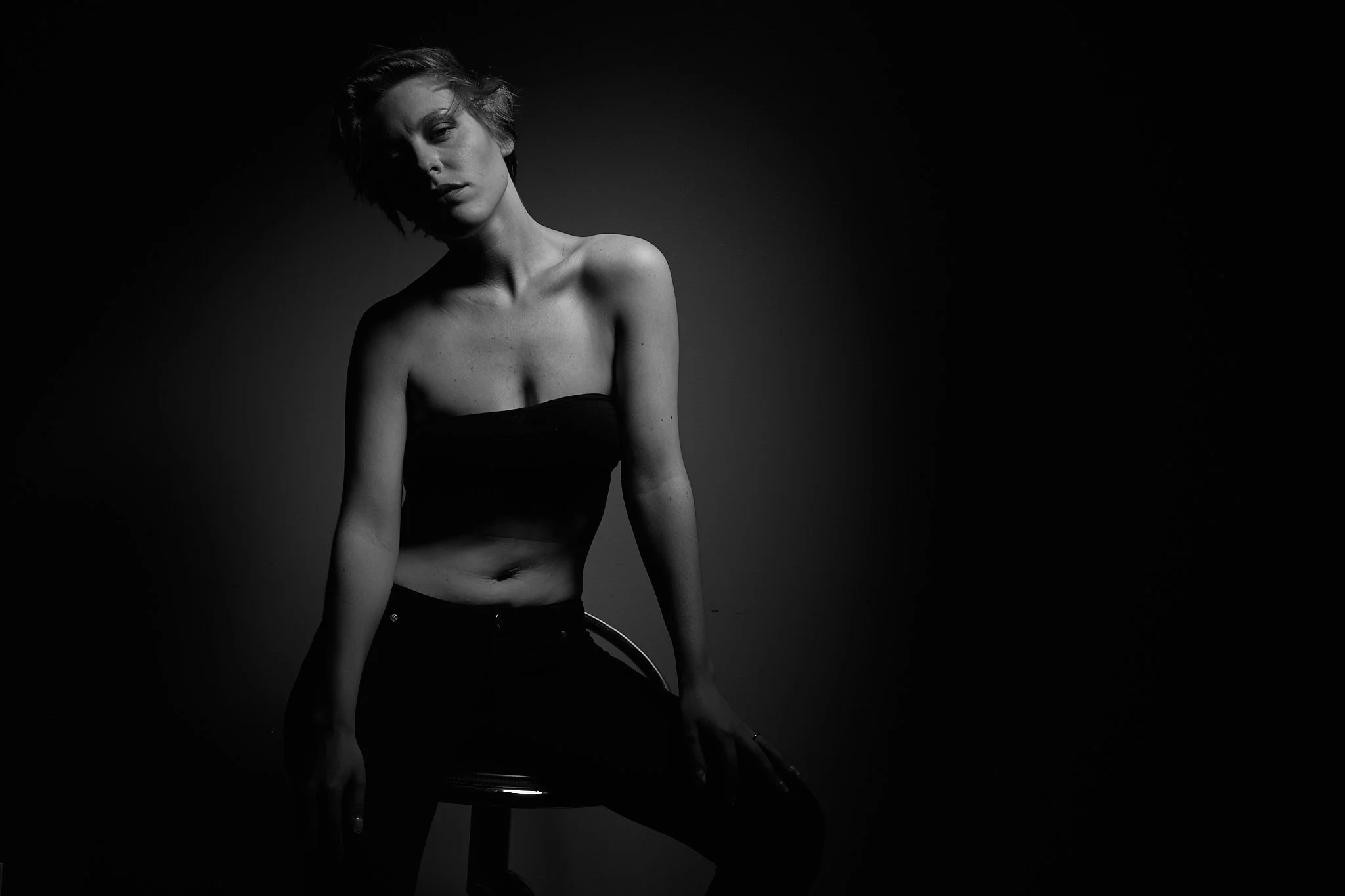 A woman with short hair sitting on a stool in a dark studio, illuminated by a single light source, wearing a strapless top and dark pants, in black and white.