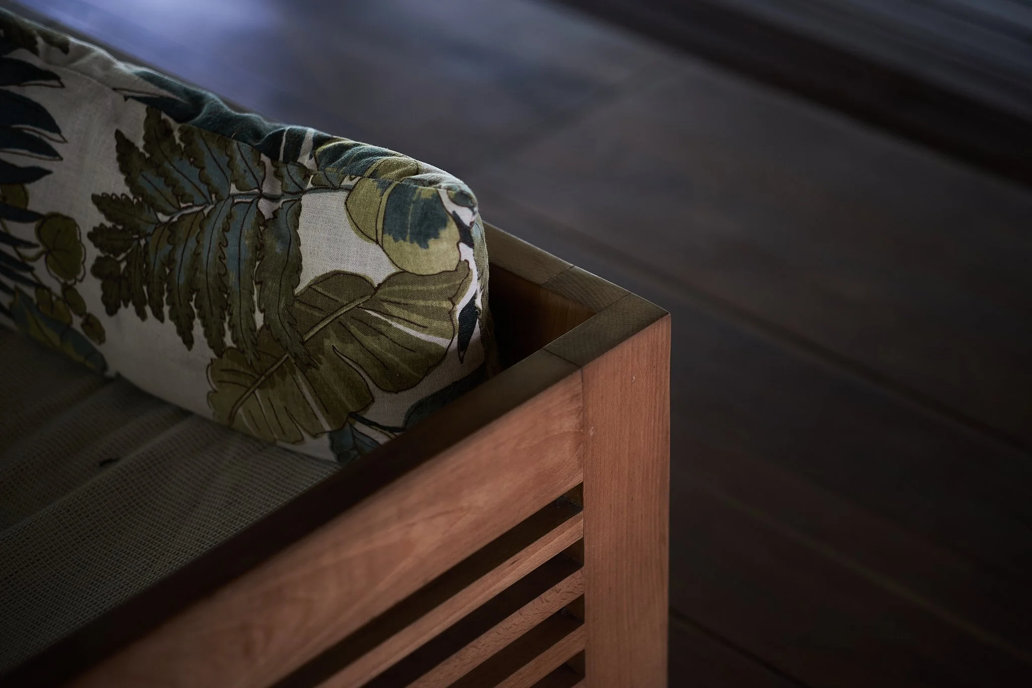 Close-up of a wooden bed frame with a tropical patterned pillow, dark wood flooring in the background