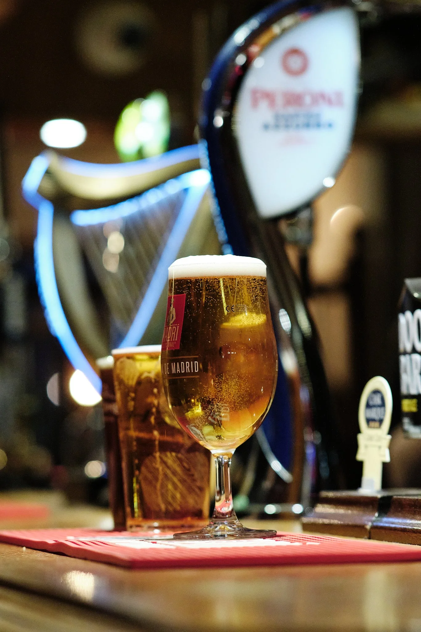 A glass of golden beer with foam on top on a bar counter, with a bottle of beer and a glass of whiskey behind it, and a blurry background of a bar setting.