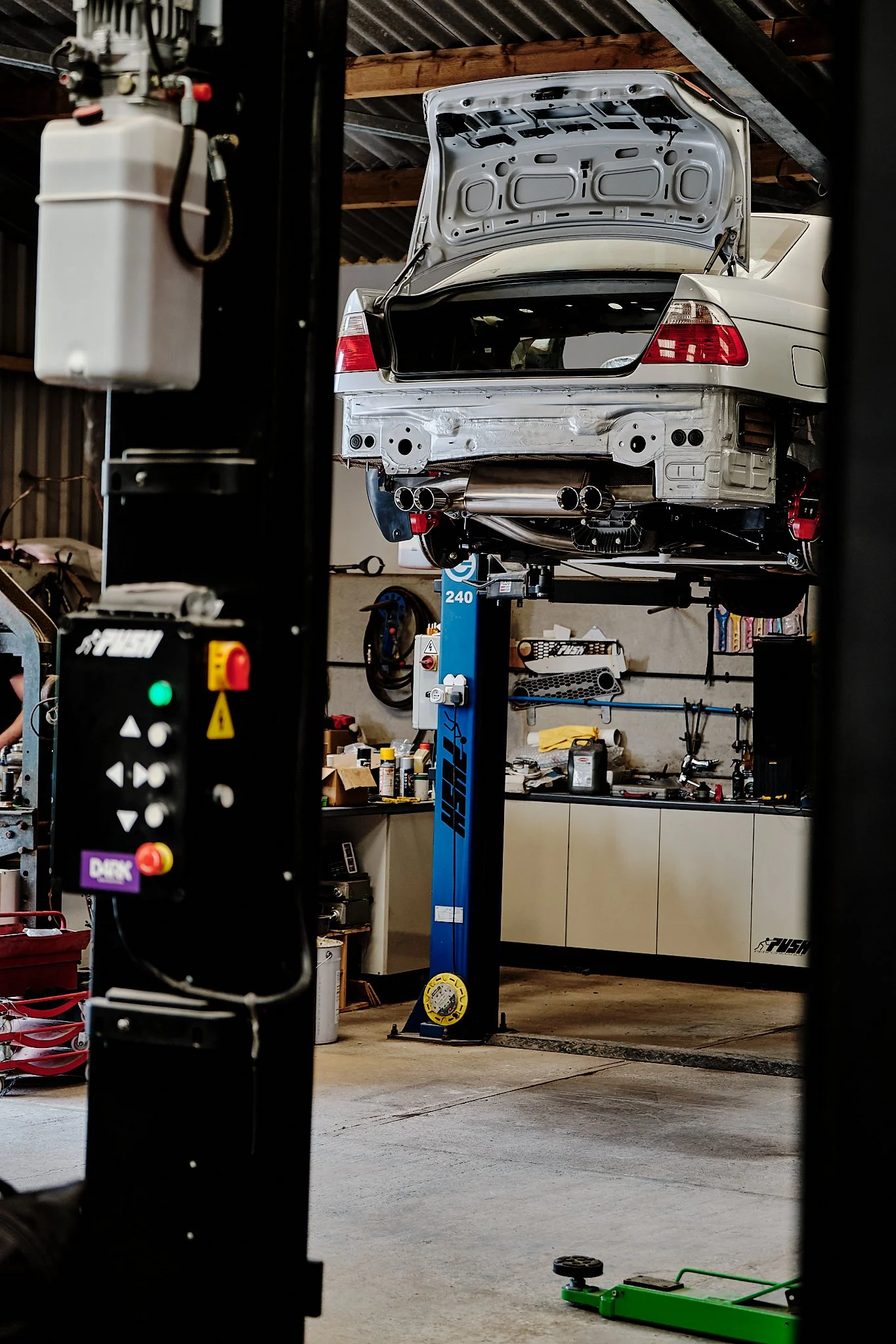 A white race car on a lift in a garage with tools and equipment. The car's rear hatch is open, exposing the trunk area, and the bumper is removed, revealing exhaust pipes.