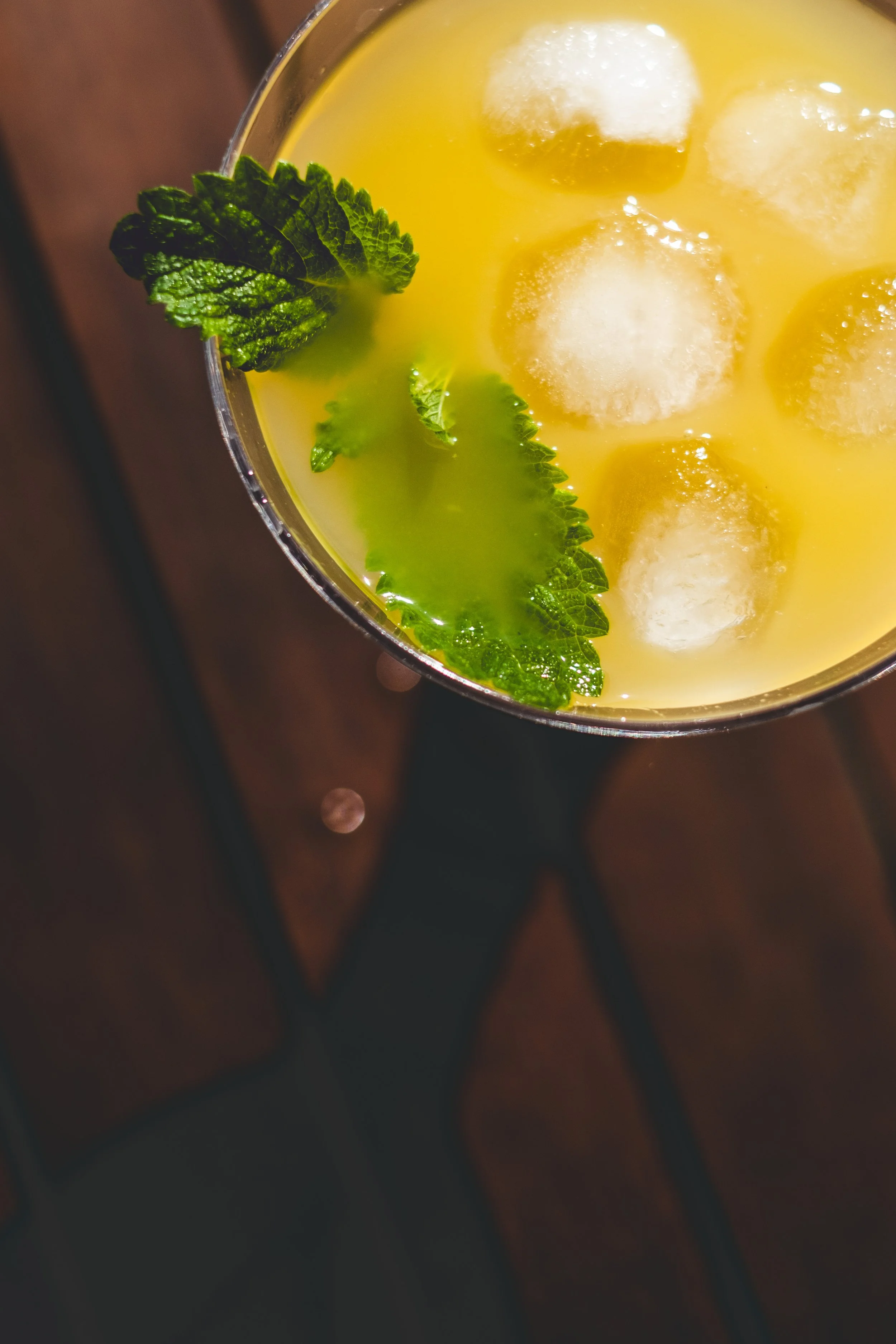 A yellow cocktail with ice cubes, garnished with a fresh mint leaf, on a dark wooden surface.