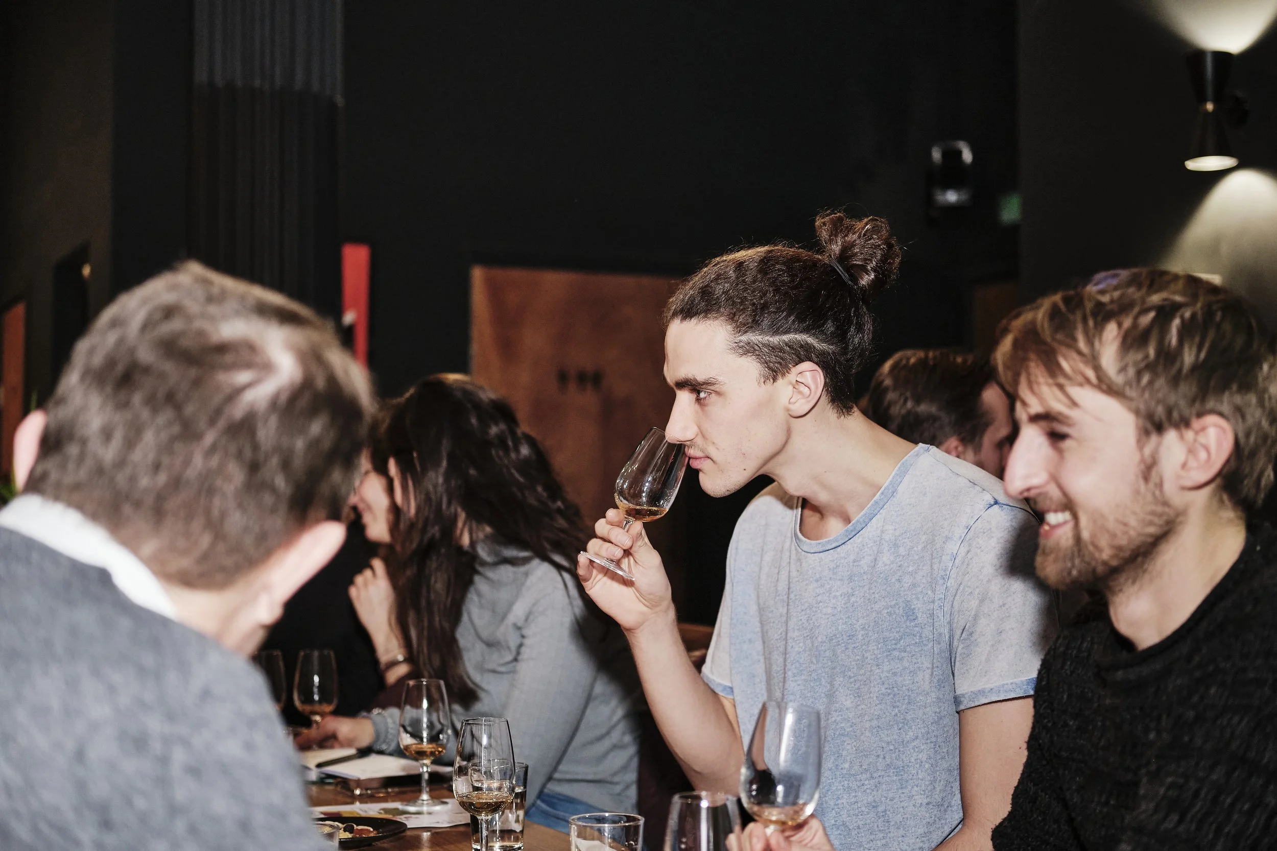 People sitting at a table in a dimly lit room, enjoying wine tasting. One person is smelling or tasting a glass of wine, smiling, while others are engaged in conversation.