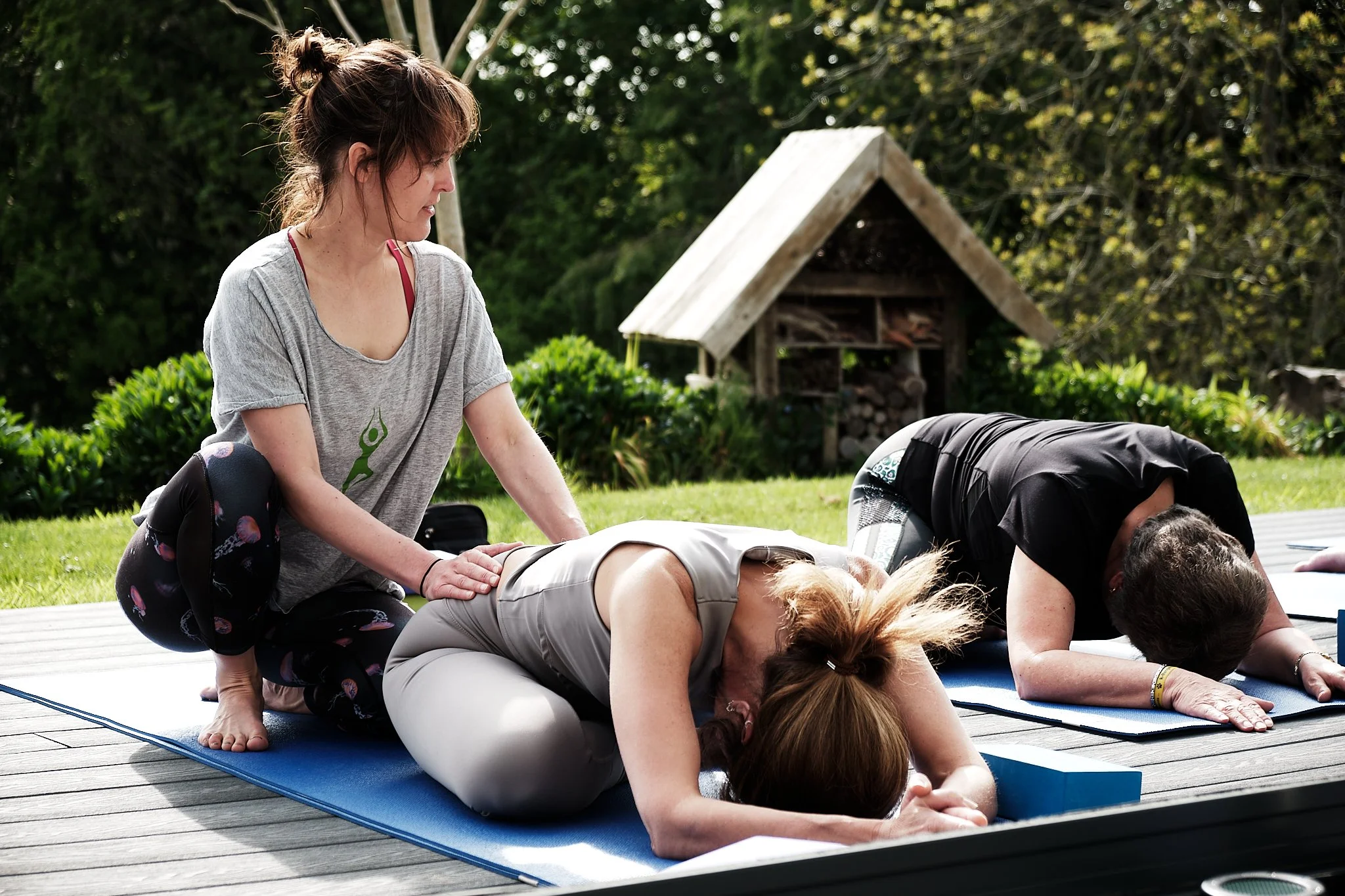 Two women practicing yoga outdoors on yoga mats, one woman assisting another with a stretching pose, on a wooden deck with greenery and a small wooden shed in the background.
