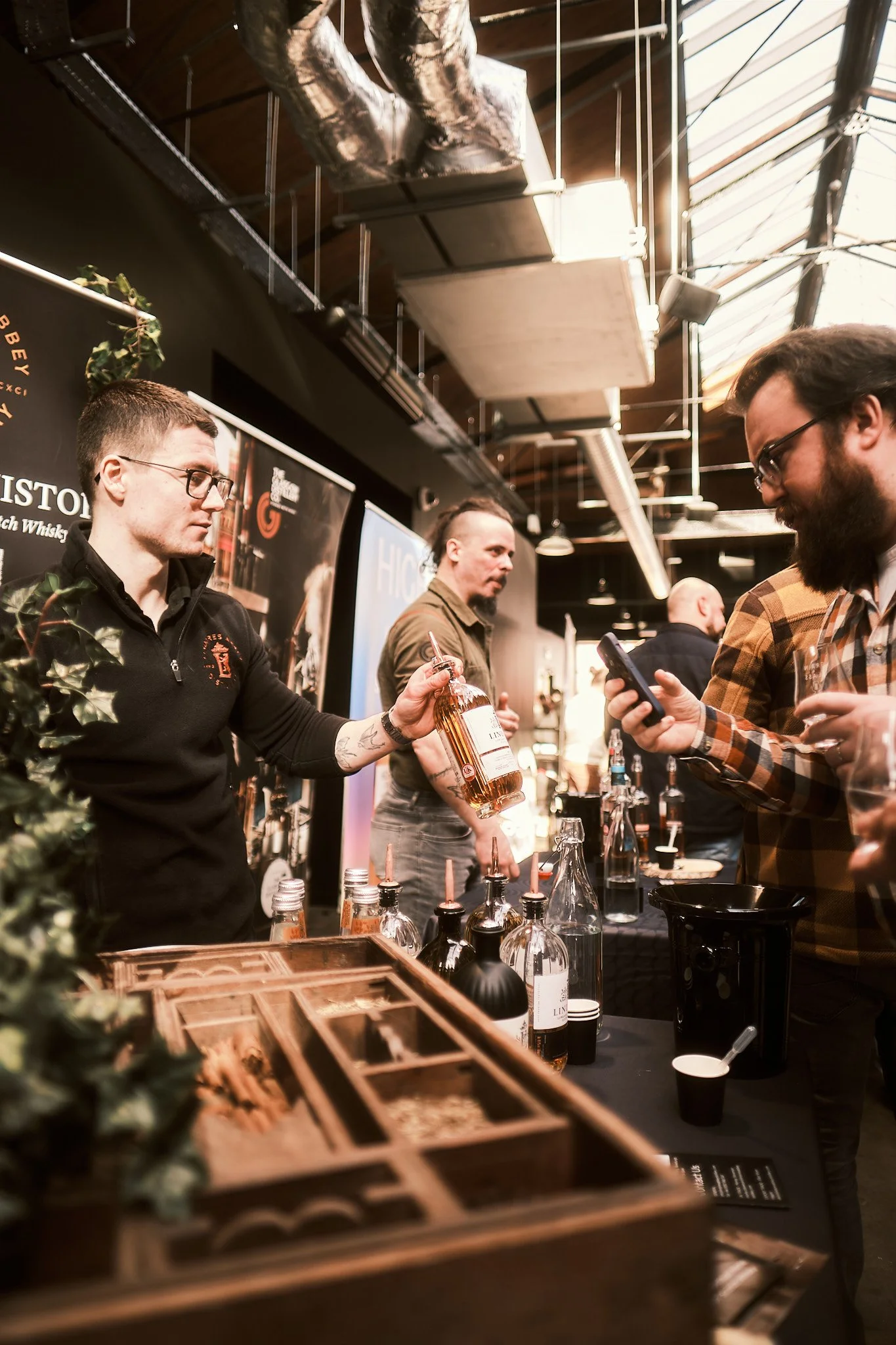 People at a whisky tasting event, with bottles of whisky on a table and a man pouring whisky, inside a venue with black walls and industrial ceiling.