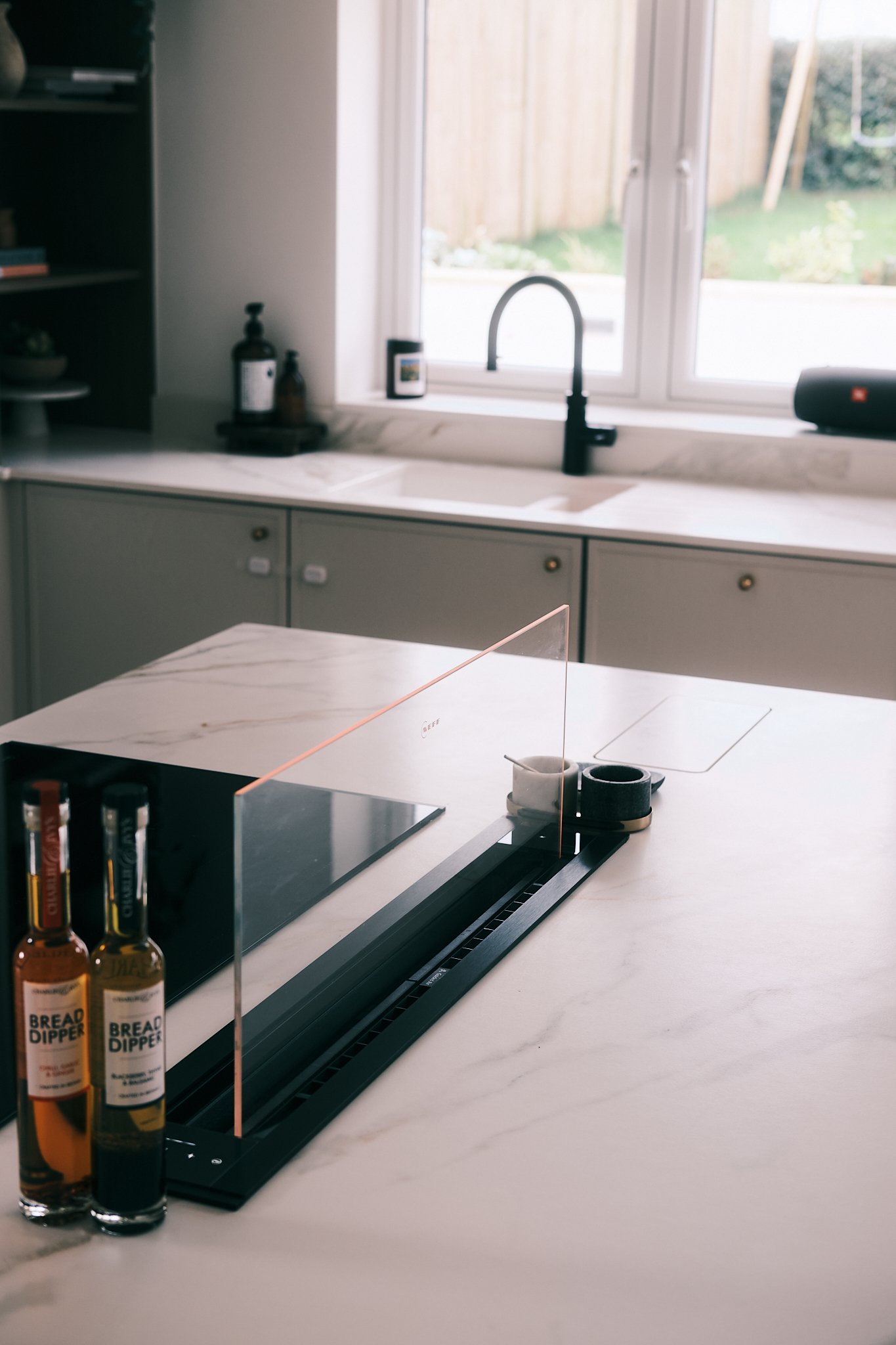 Modern kitchen with white marble island, black sink, and a window with a view of a wooden fence and greenery.
