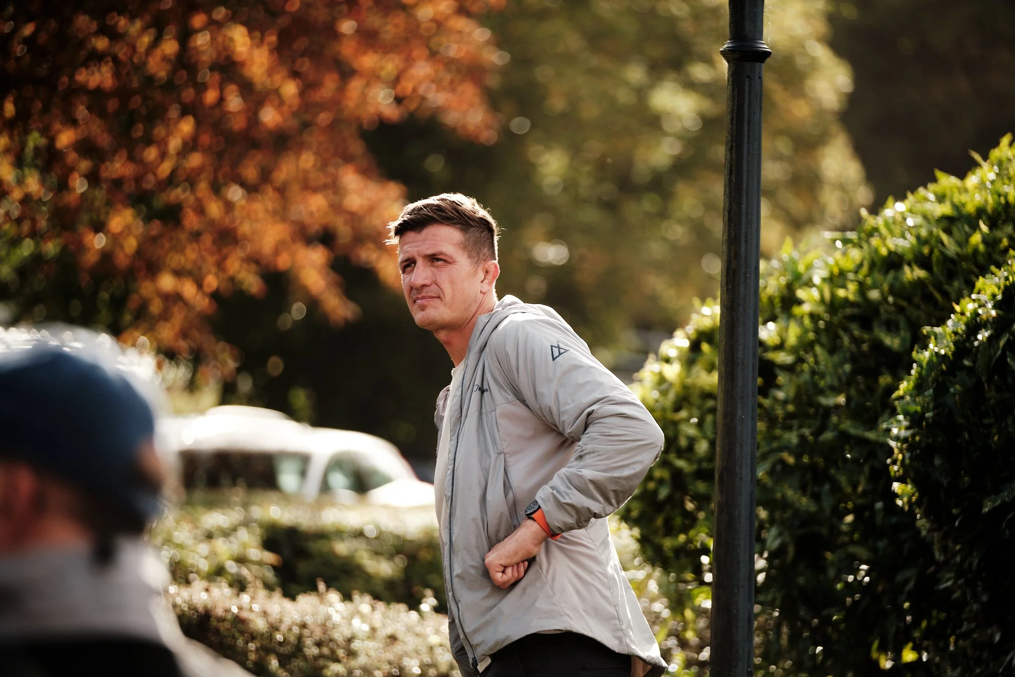 A man in a gray jacket stands outdoors with his hands on his hips, looking over his shoulder, in a setting with autumn trees and a car in the background.