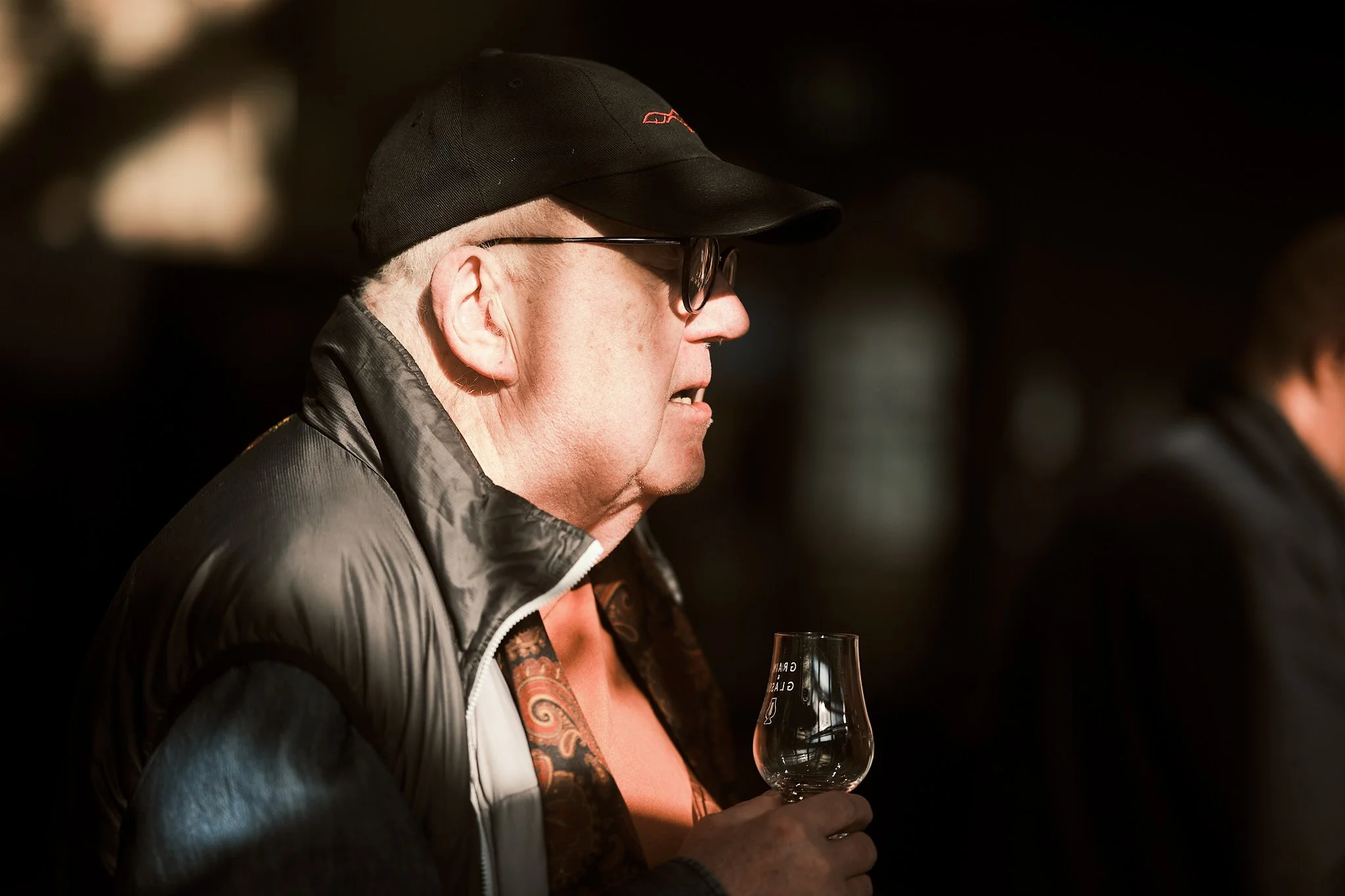 Older man wearing glasses, a black cap, and a black jacket, holding a glass of whiskey at an event.