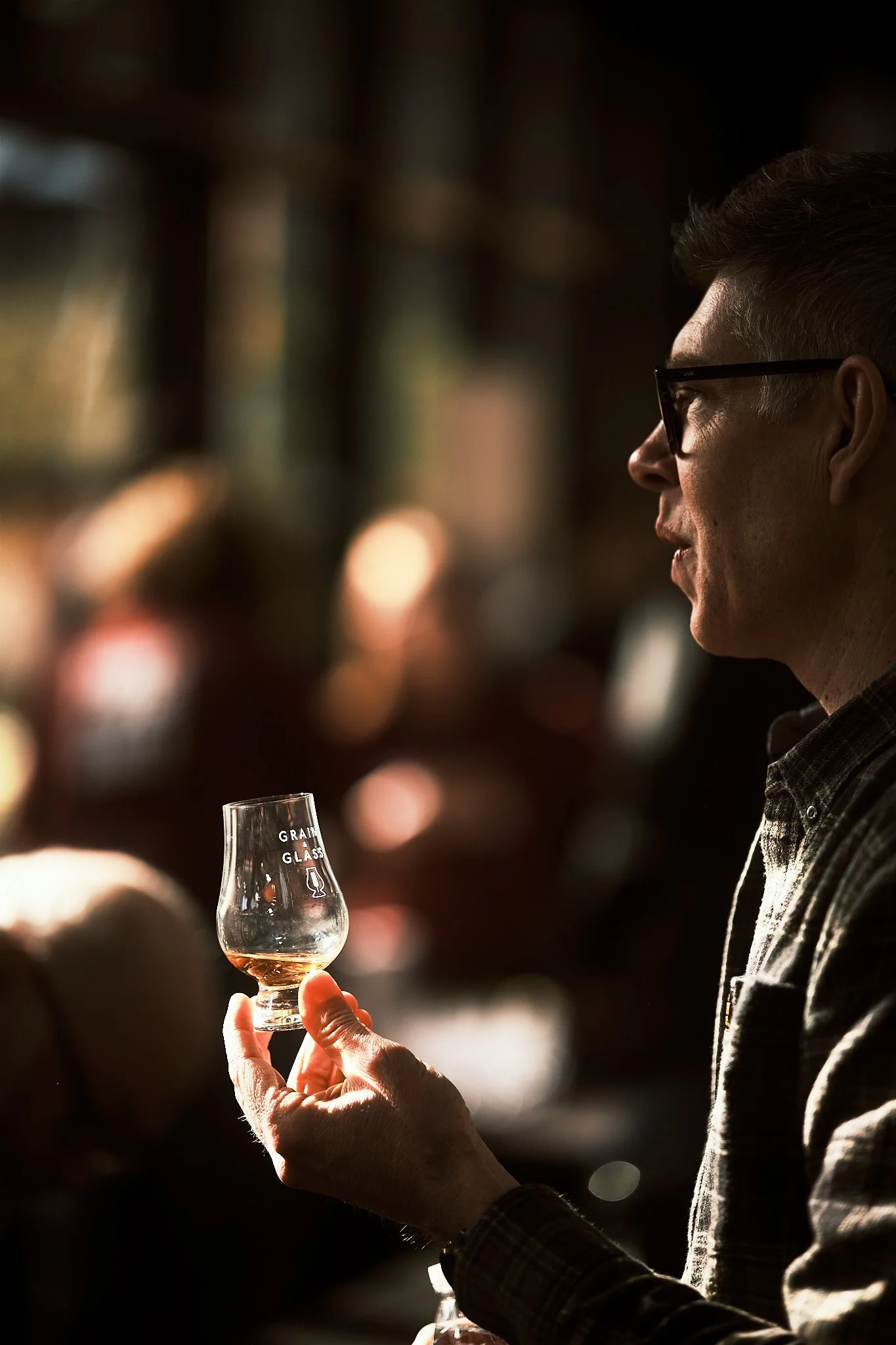 A man with glasses holding a small glass of whiskey in a dimly lit bar with blurred background.