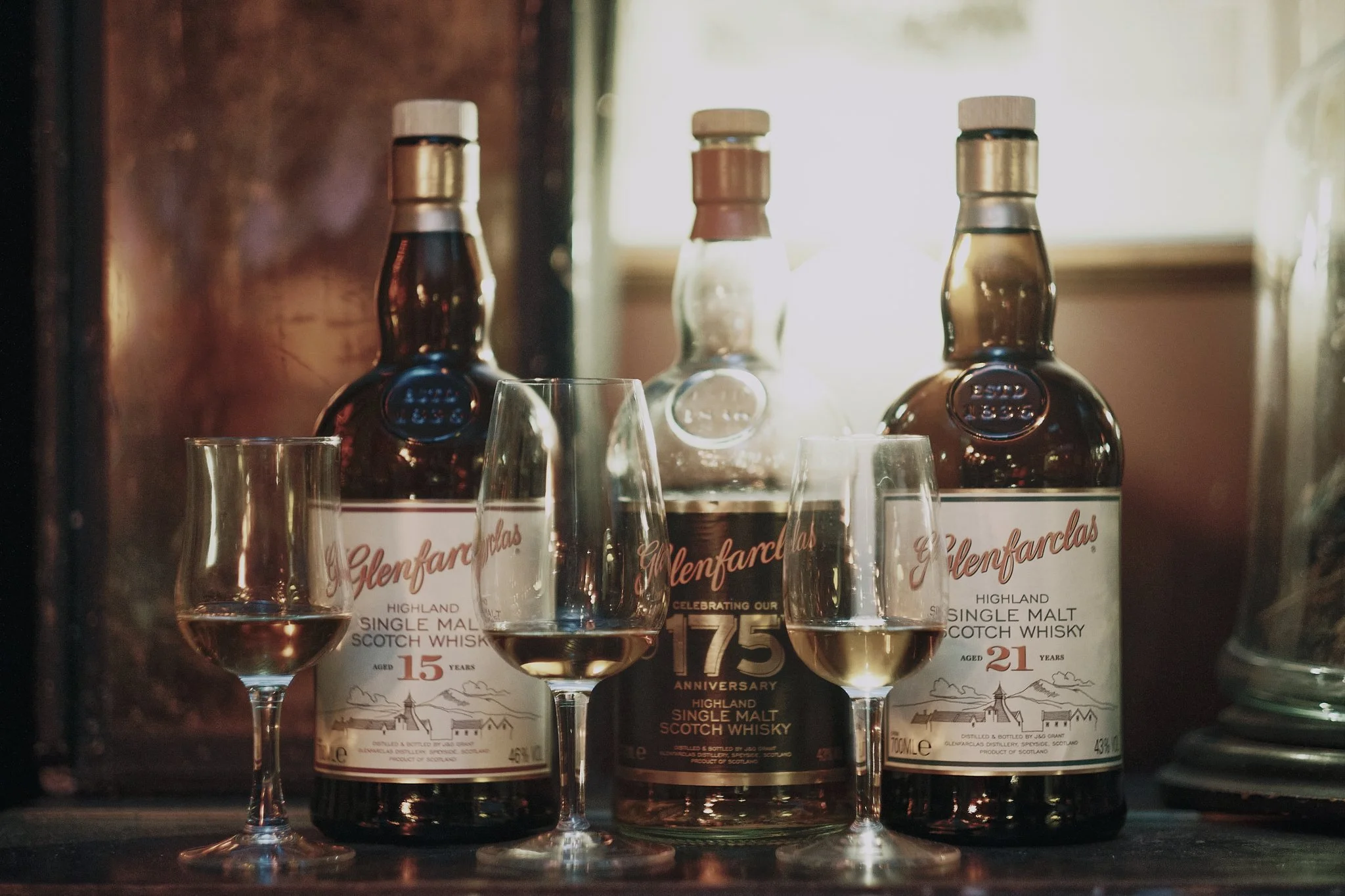 Three bottles of Glenfarclas Scotch whisky and three glasses filled with whisky on a wooden surface, with a backlit window in the background.