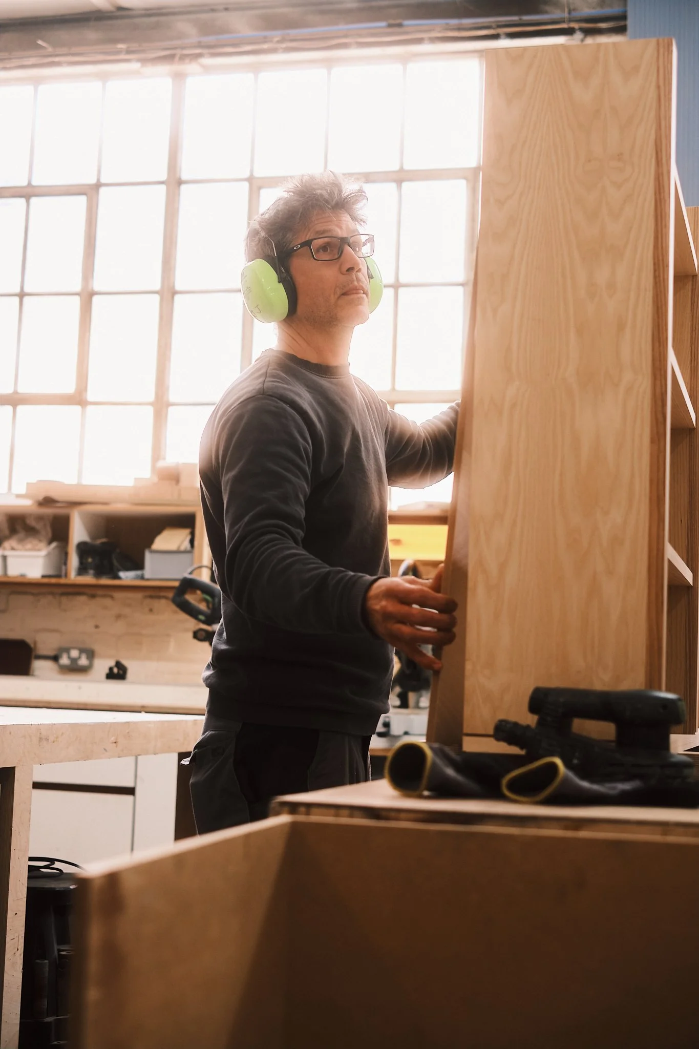 Man working on a woodworking project in a workshop, wearing green ear protection and glasses, with wooden furniture and tools around.