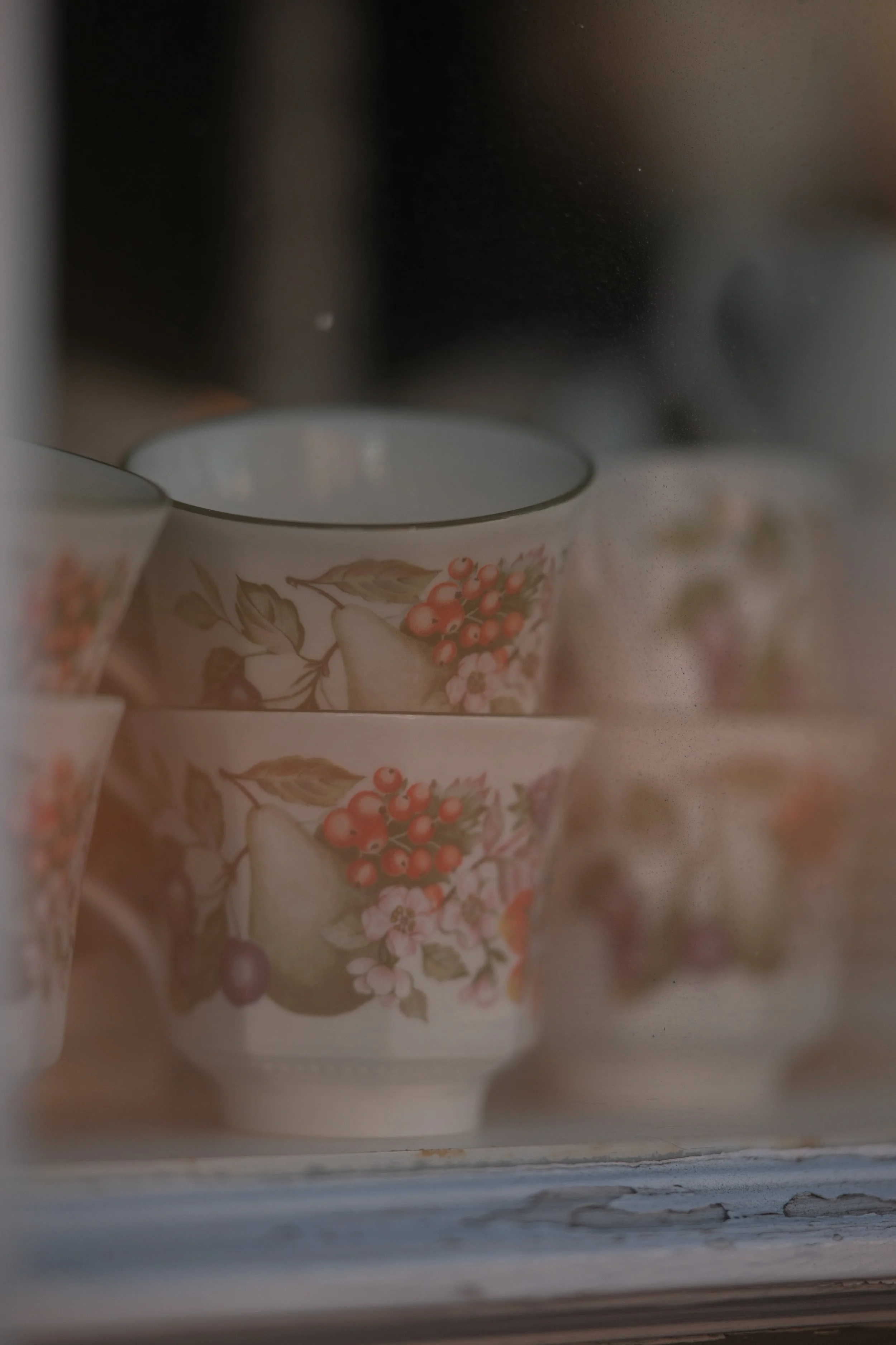 Stacked floral patterned ceramic cups with fruit and flowers, seen through a slightly foggy glass door.