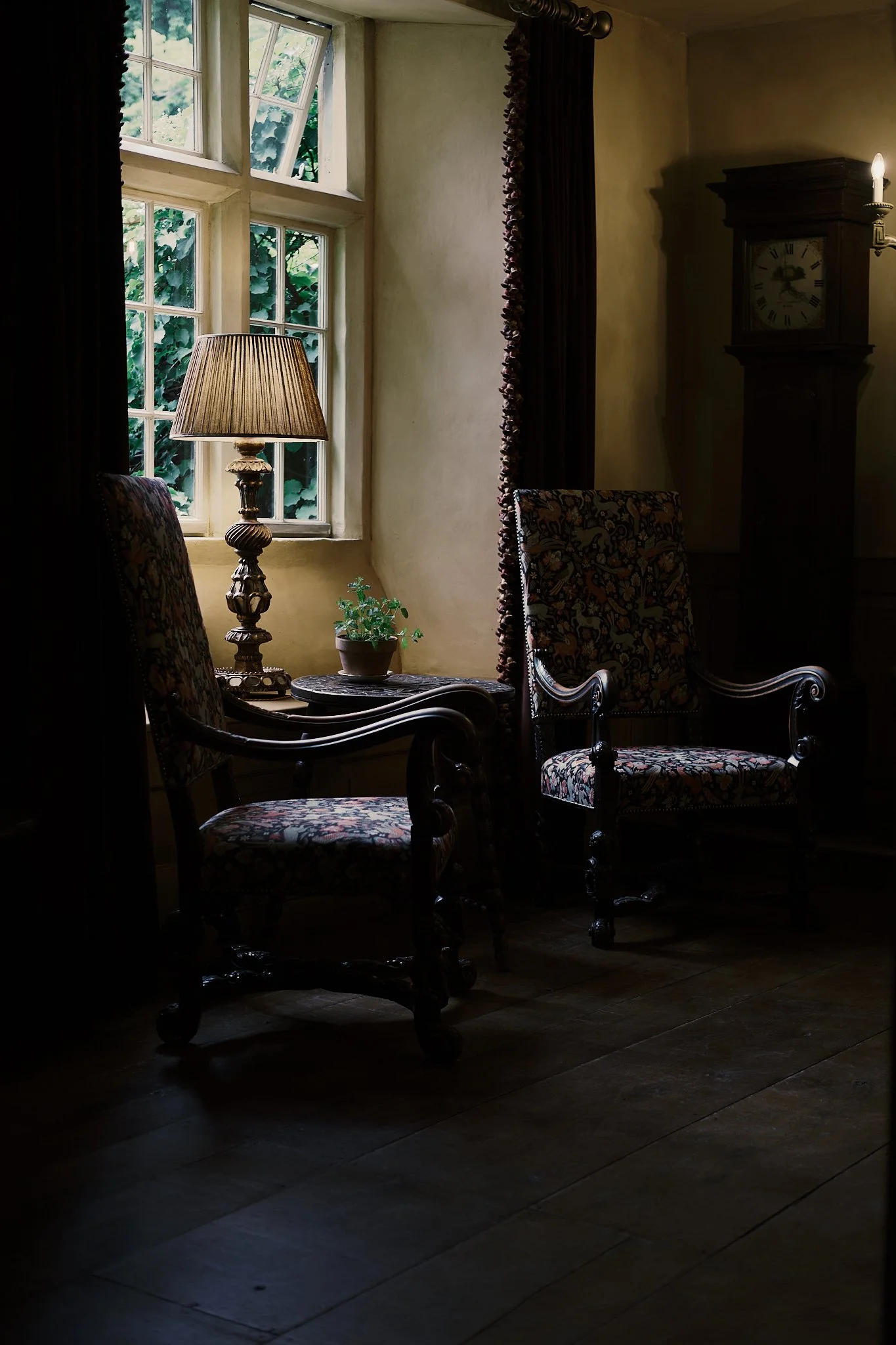 A cozy corner in a vintage room with two floral upholstered armchairs beside a small table holding a potted plant and a lamp. There are dark curtains and a tall grandfather clock near a window that lets in natural light, illuminating part of the room
