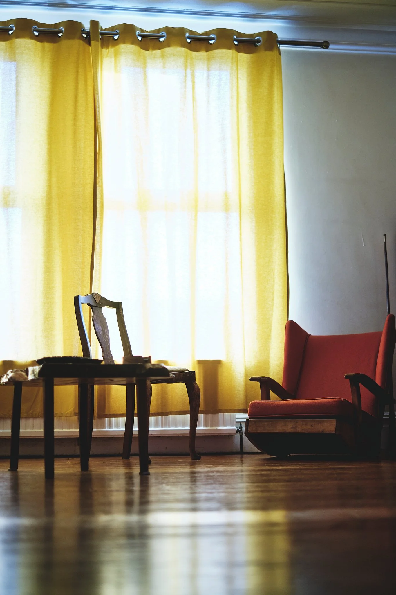 A room with yellow curtains, a vintage wooden chair, and a red upholstered armchair, with sunlight filtering through the curtains onto a hardwood floor.