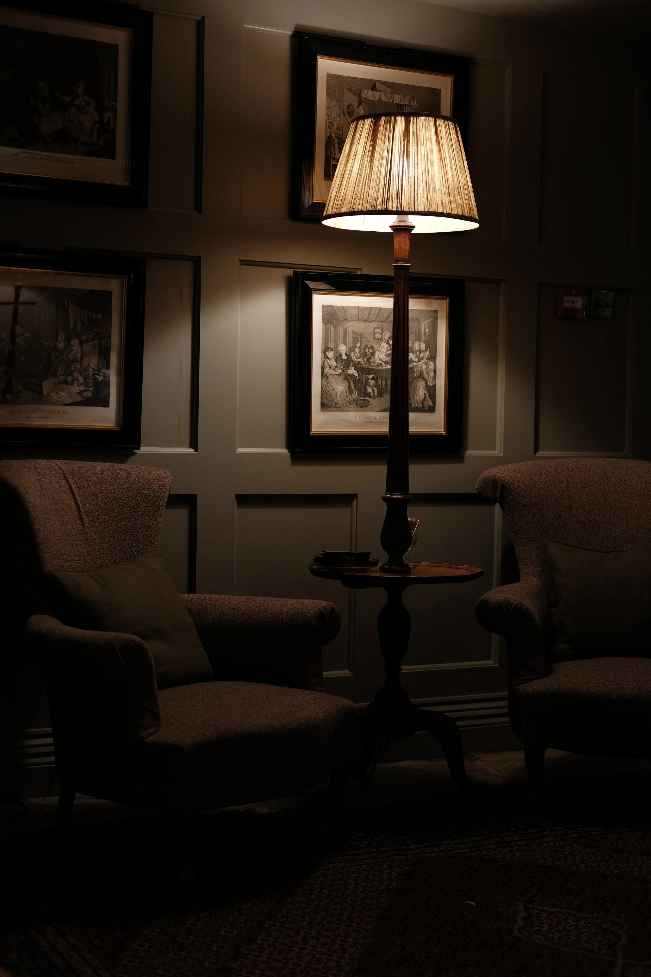 Dimly lit sitting area with two armchairs, a side table, and a tall floor lamp with a beige shade. Framed artwork decorates the wall behind, and the carpet is visible on the floor.