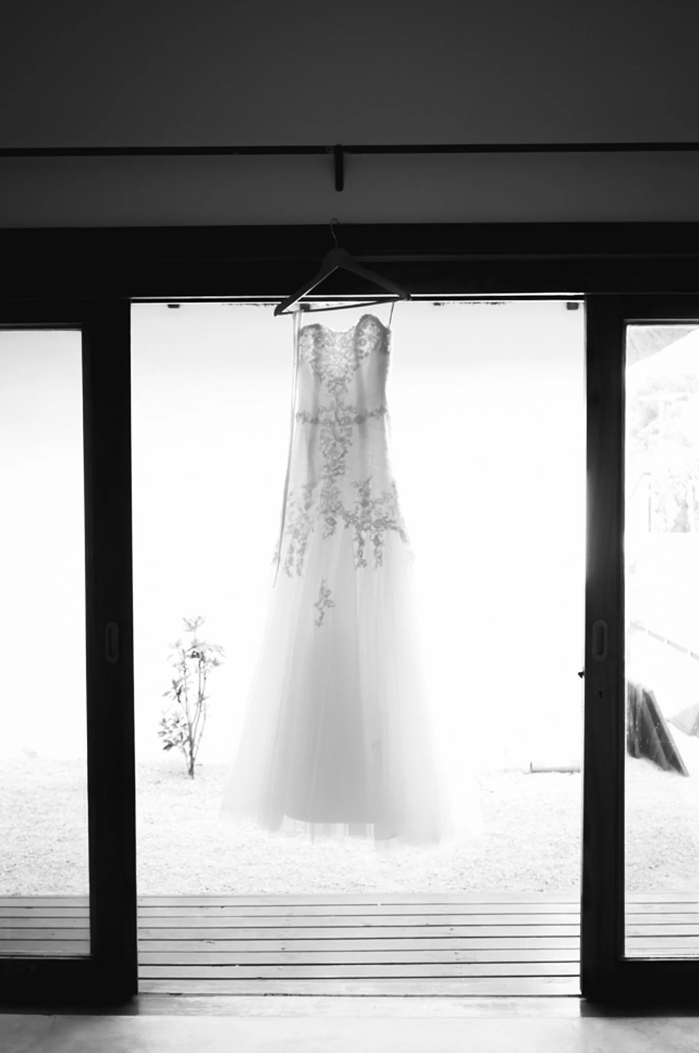 Black and white photo of a wedding dress hanging on a hanger in front of large windows, with a landscape visible outside.