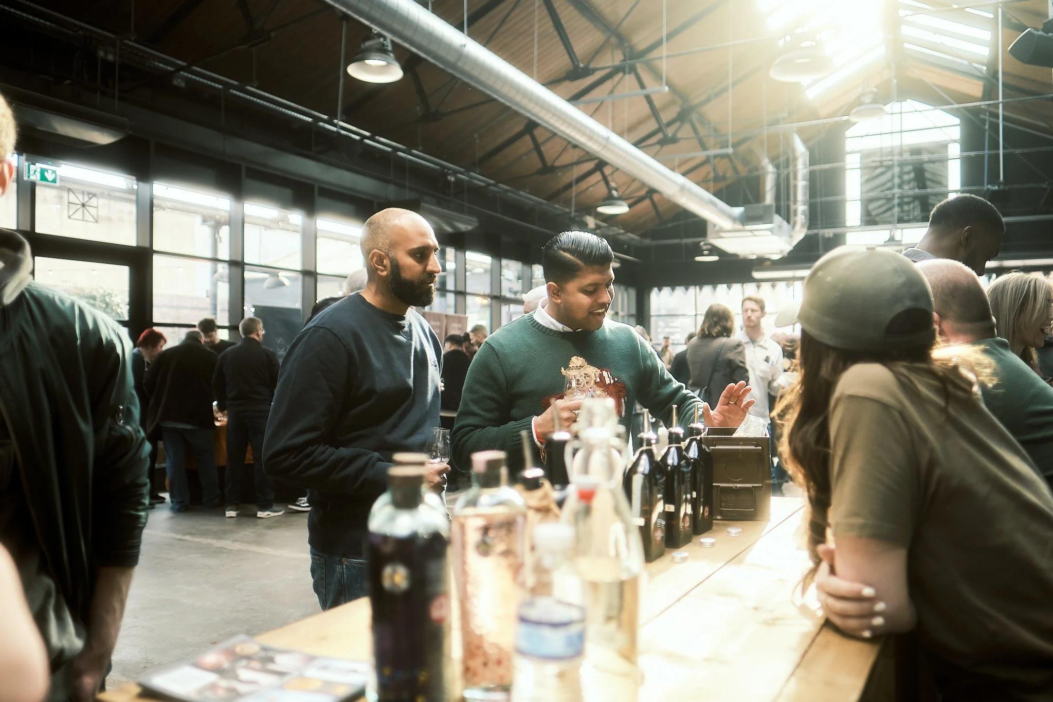 People socializing at a bar or cafe with large windows and industrial ceiling in a bright, spacious setting.