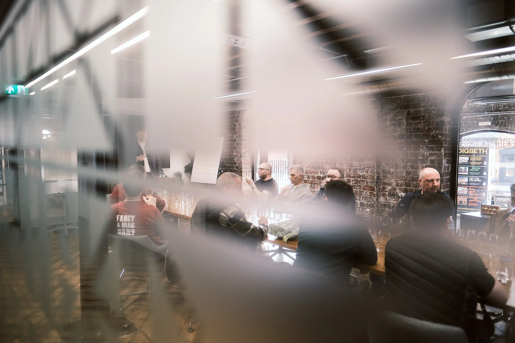 People gathered around a long table in a contemporary meeting room with exposed brick walls, seen through a blurred glass or partition.
