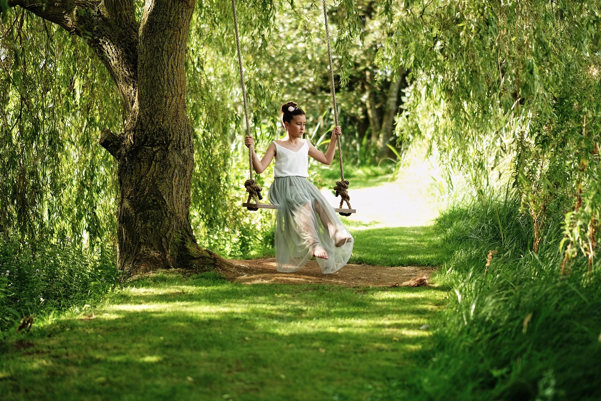 A young girl dressed in a white top and flowing skirt sitting on a swing hanging from a large tree in a lush green park with sunlight filtering through the leaves.