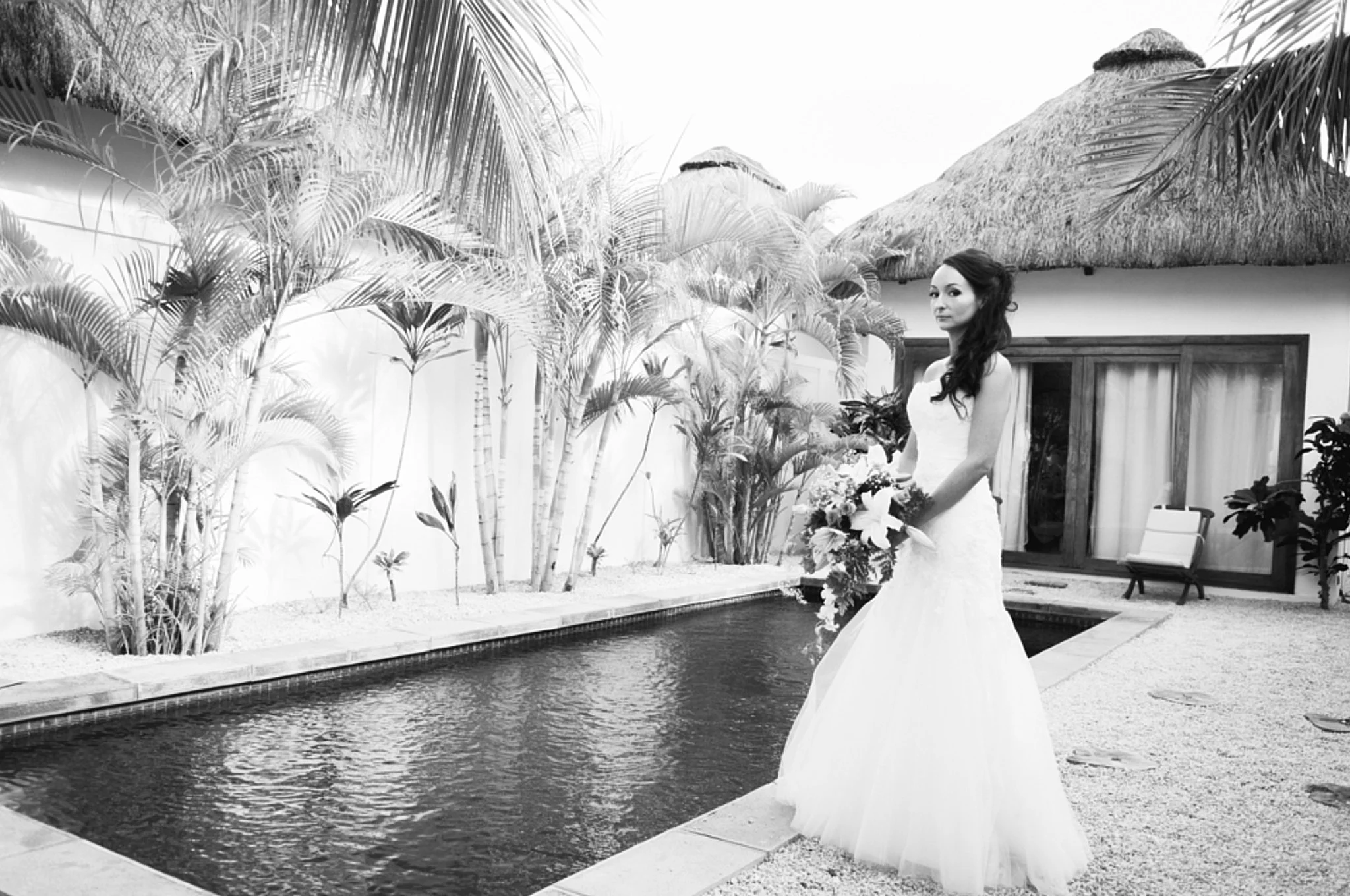 A woman in a strapless wedding dress holding a bouquet of flowers, standing near a pool in a tropical outdoor setting with palm trees and a thatched roof hut.