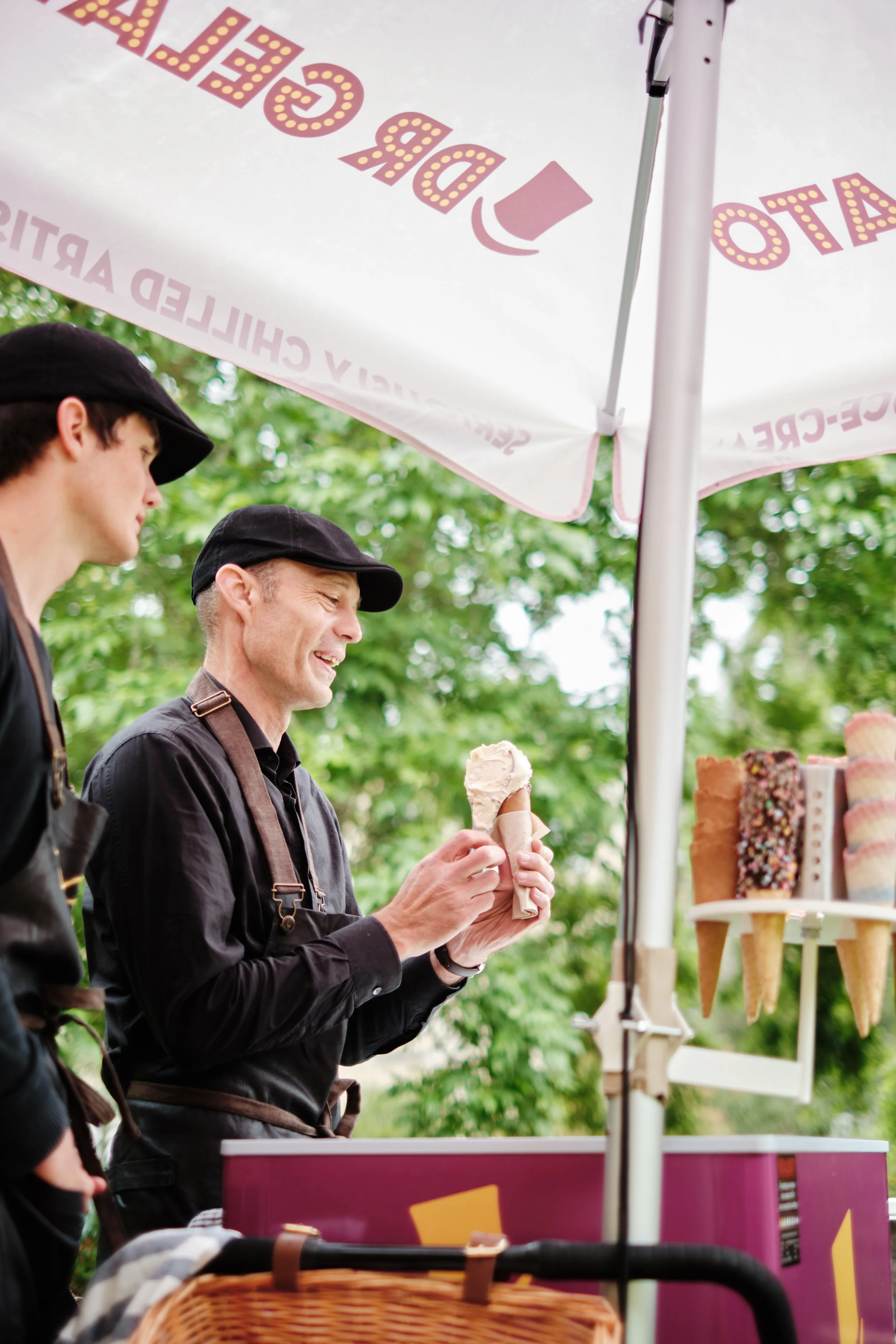 A man at an outdoor ice cream stand holding a cone with a scoop of vanilla ice cream under a large umbrella with the text 'STATO' and 'CHILLY CHARITIES'. Two other people are partially visible next to him.