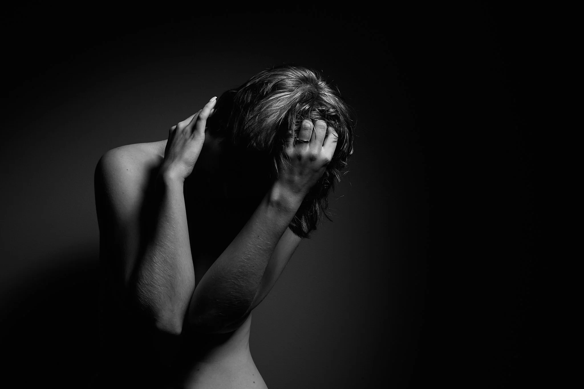 Black and white photo of a woman with short hair, covering her face with her hands, against a dark background.