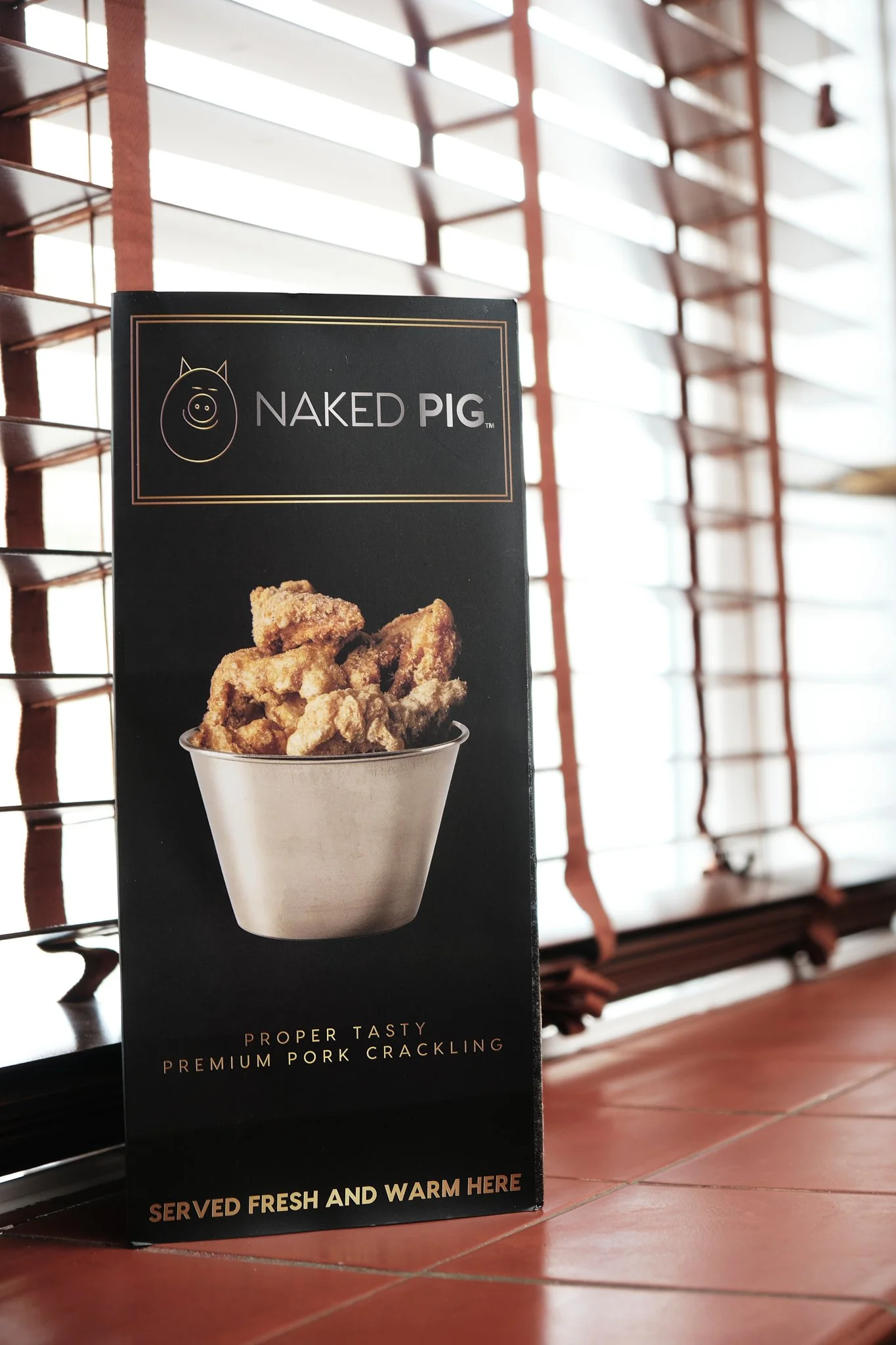 Signboard for Naked Pig showing an image of fried pork cracklings in a bowl with the text "Proper Tasty Premium Pork Crackling" and "Served Fresh and Warm Here".