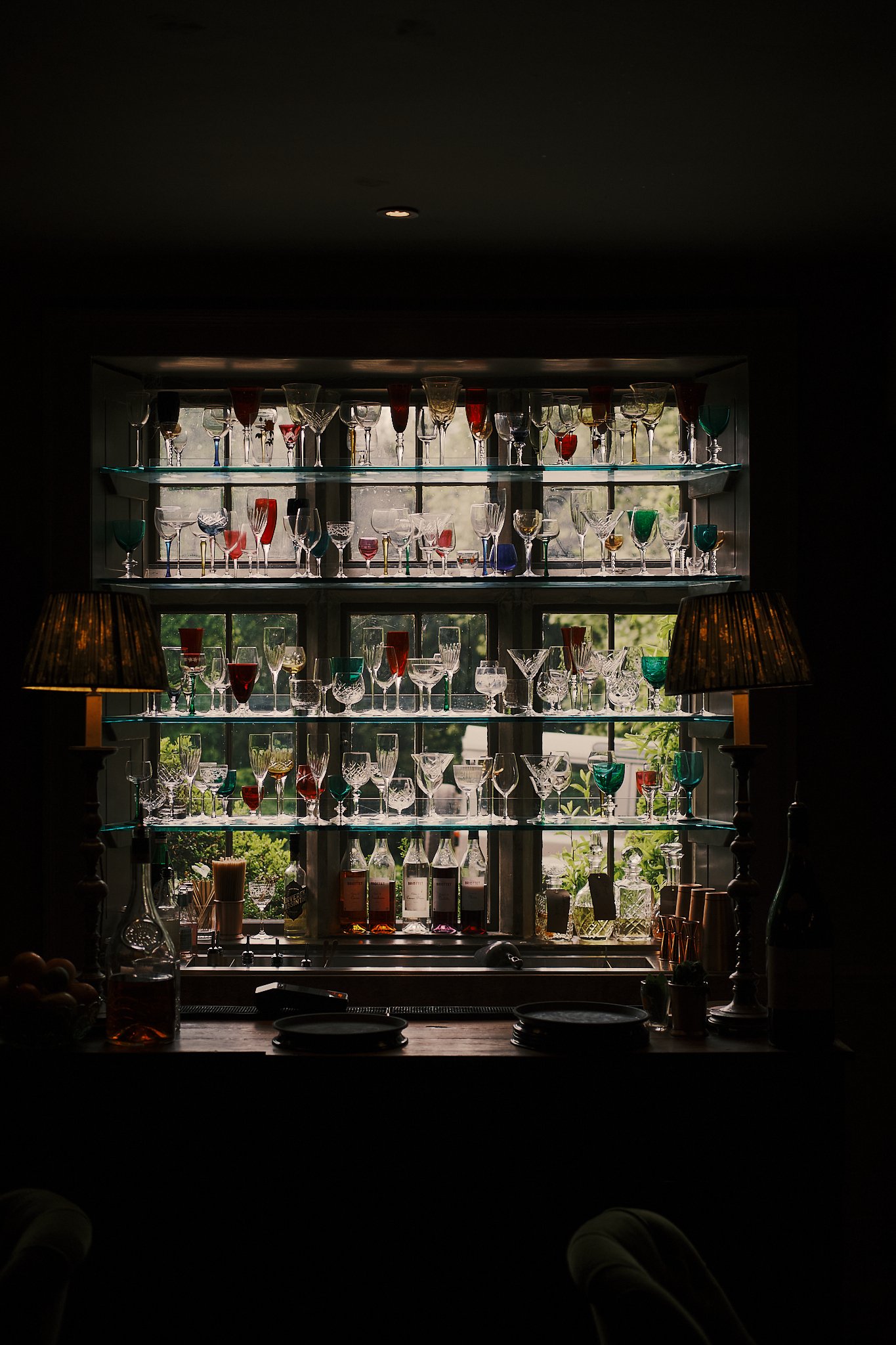 Display of colorful glassware on glass shelves in front of a window, with indoor lighting and a dark interior. Hotel interior photography at THE PIG-in the Cotswolds