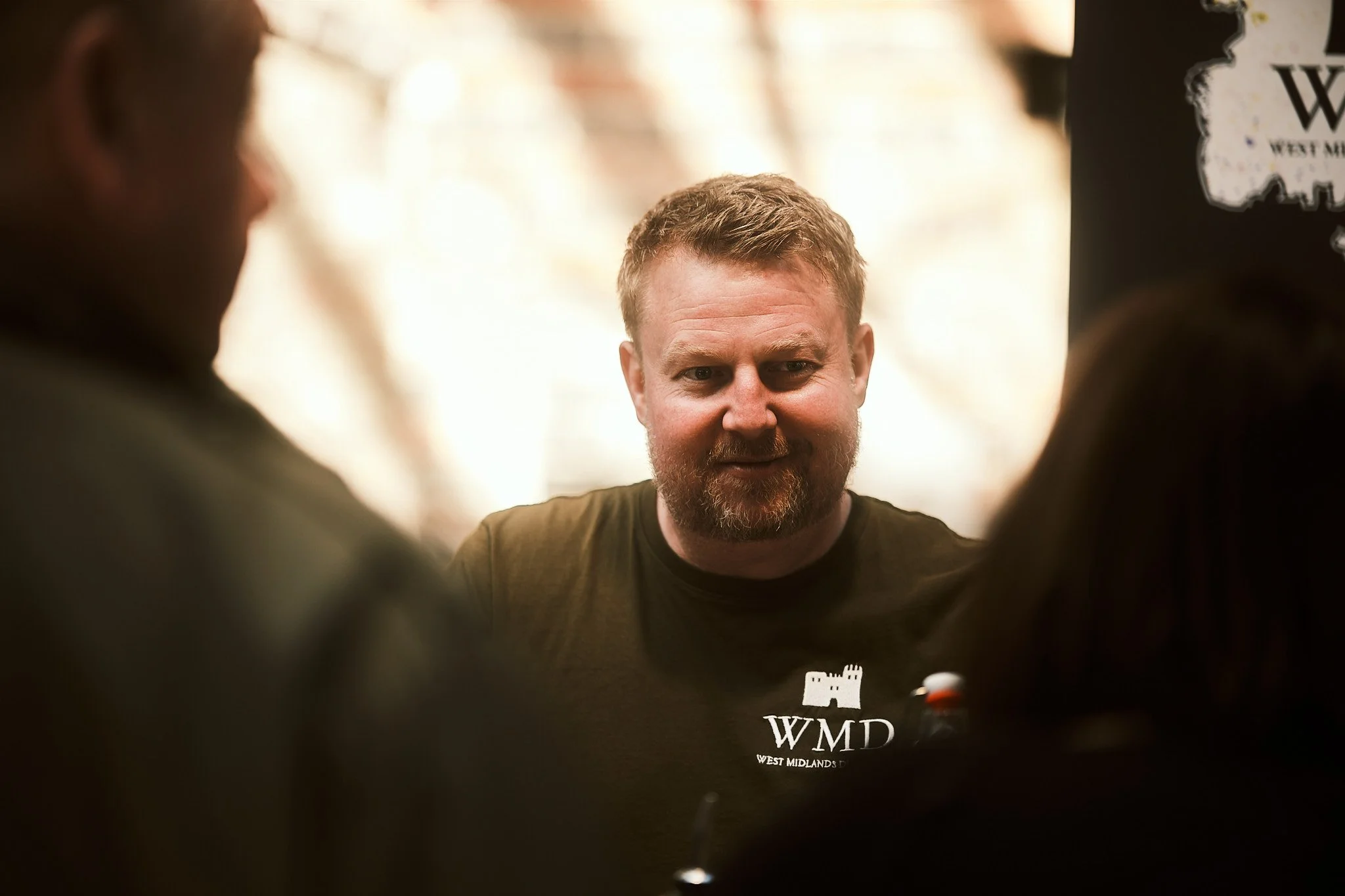 A man with a beard and short hair, wearing a black T-shirt with 'WMD West Midlands' text and a castle logo, sitting indoors with blurred people in the foreground.