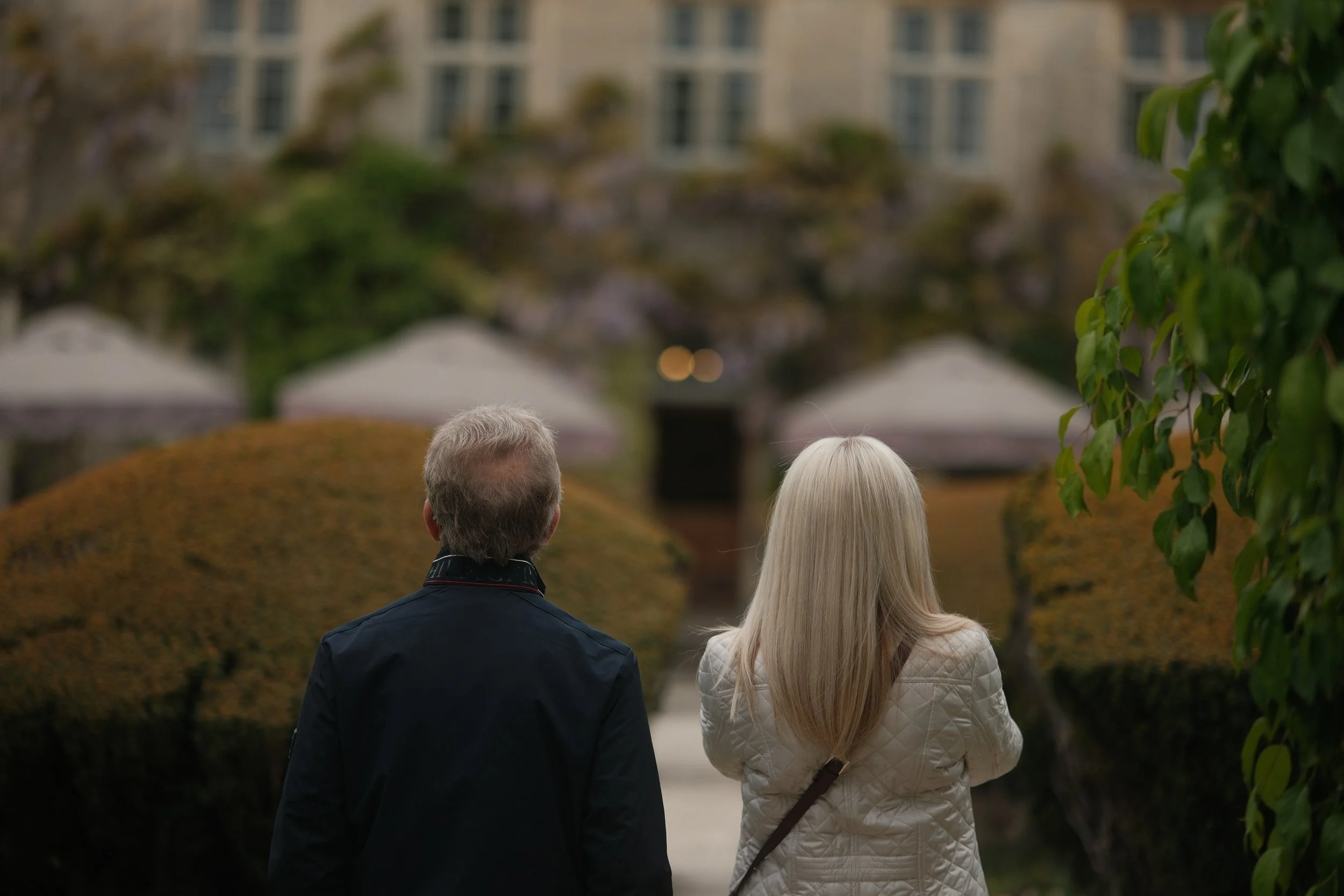 A man and a woman with light hair walking together in an outdoor park or garden, viewed from behind, with trees and bushes around.