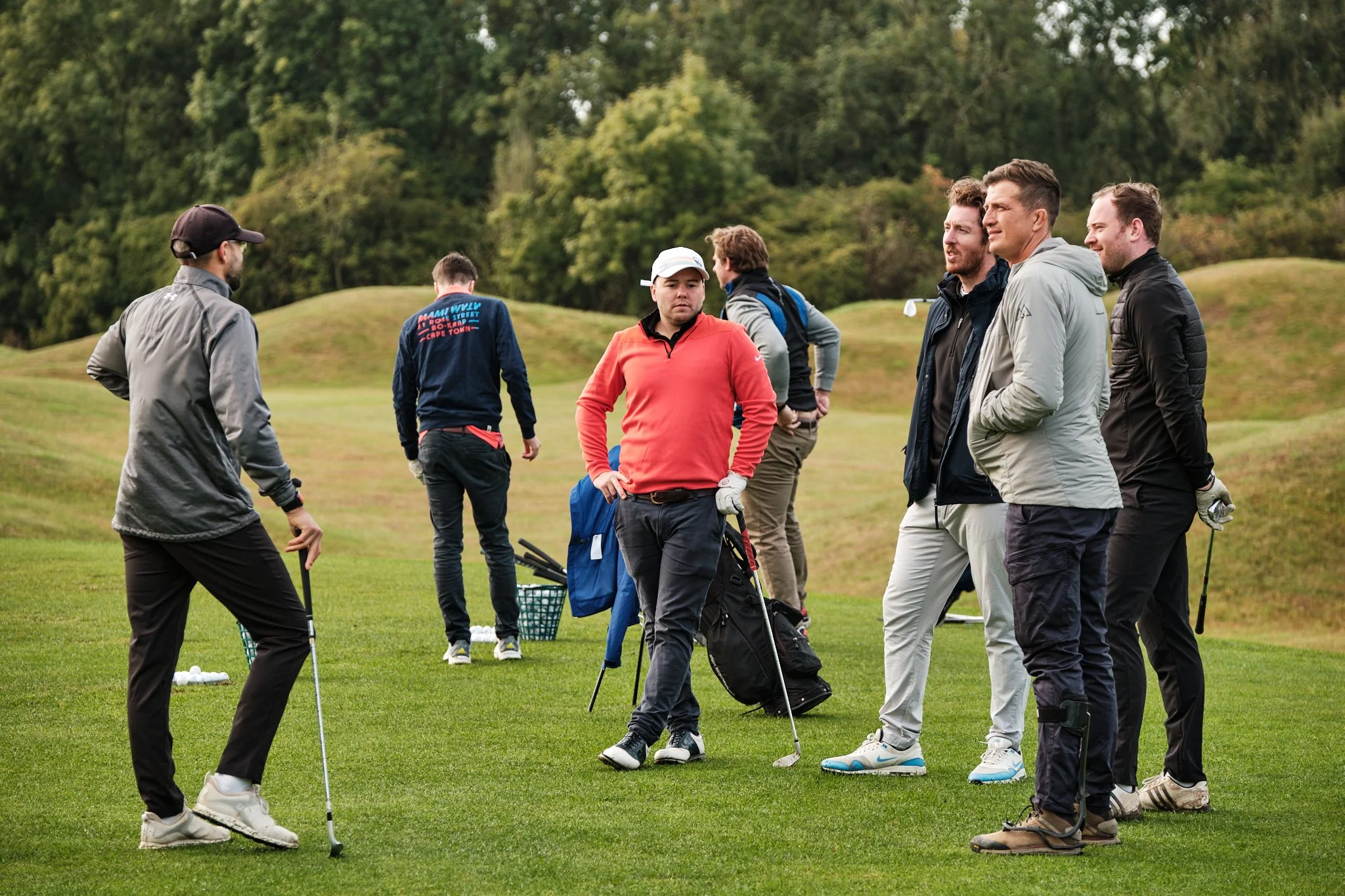 A group of men on a golf course, some holding golf clubs and standing on the green with hilly terrain and trees in the background.