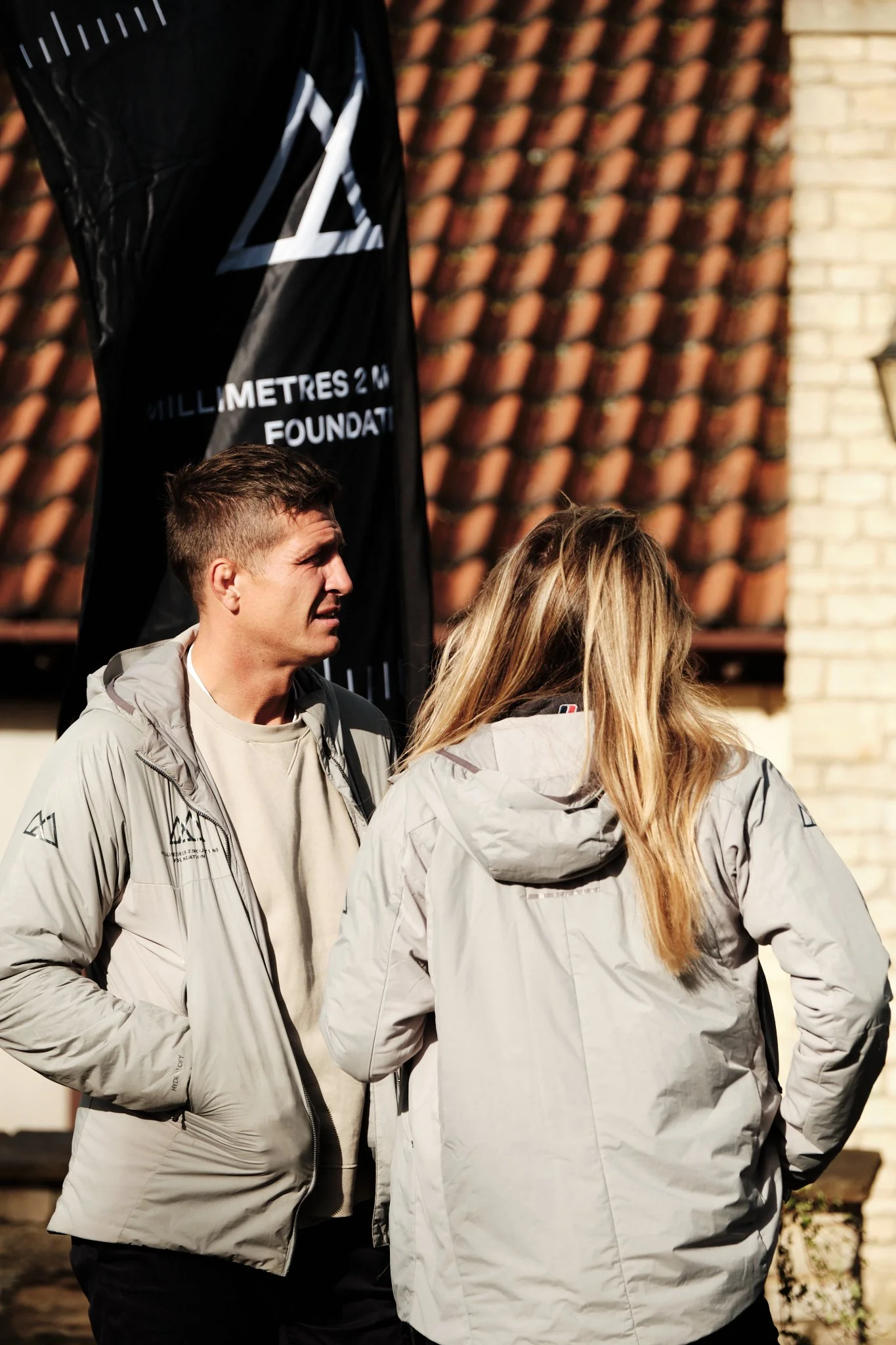 Two people in light-colored jackets conversing outdoors near a black banner with text and a house with a tiled roof in the background.