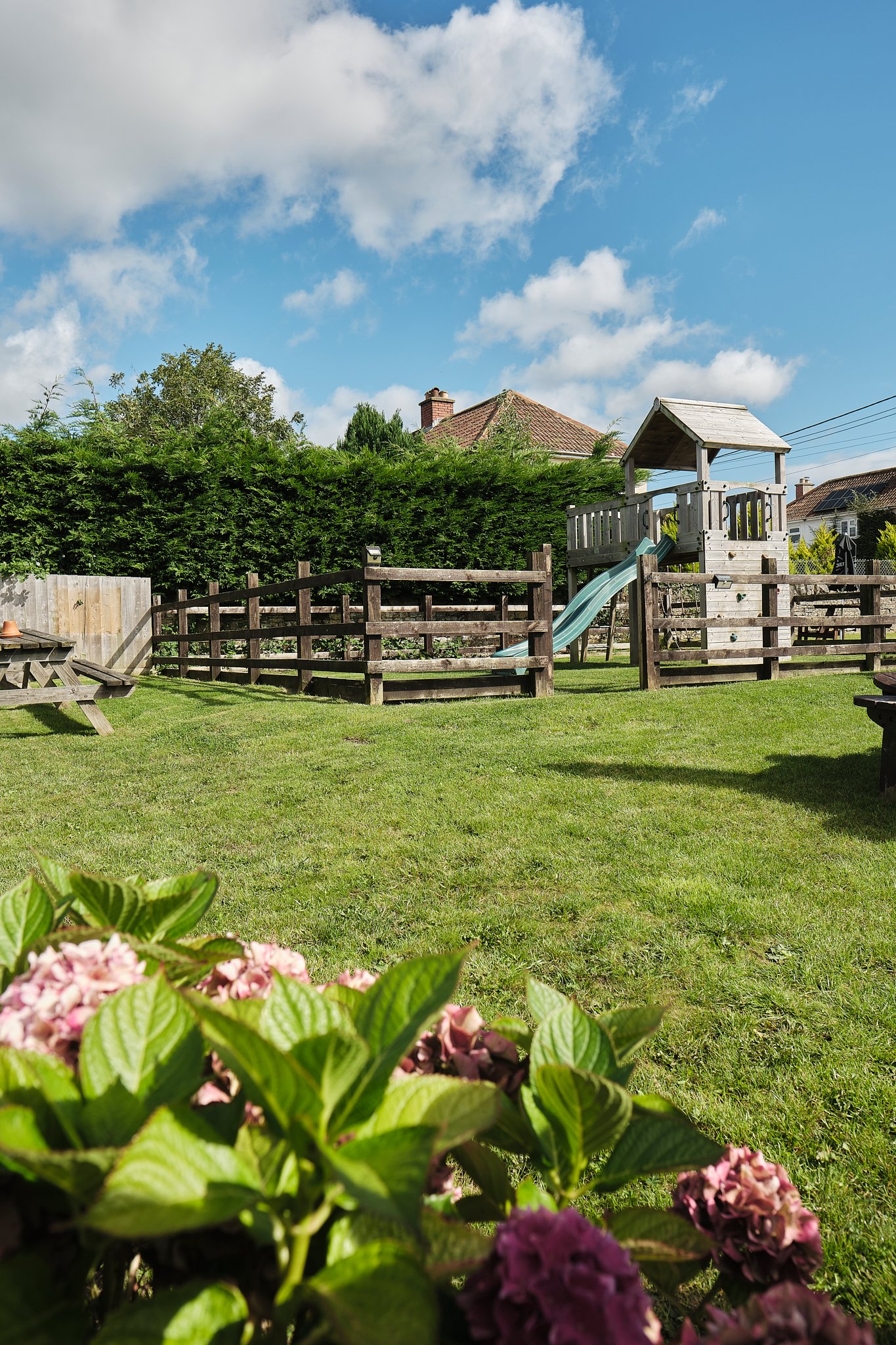 A backyard with a well-maintained lawn, a wooden play structure with a slide, surrounded by a wooden fence, pink flowers in the foreground, and a partly cloudy sky.