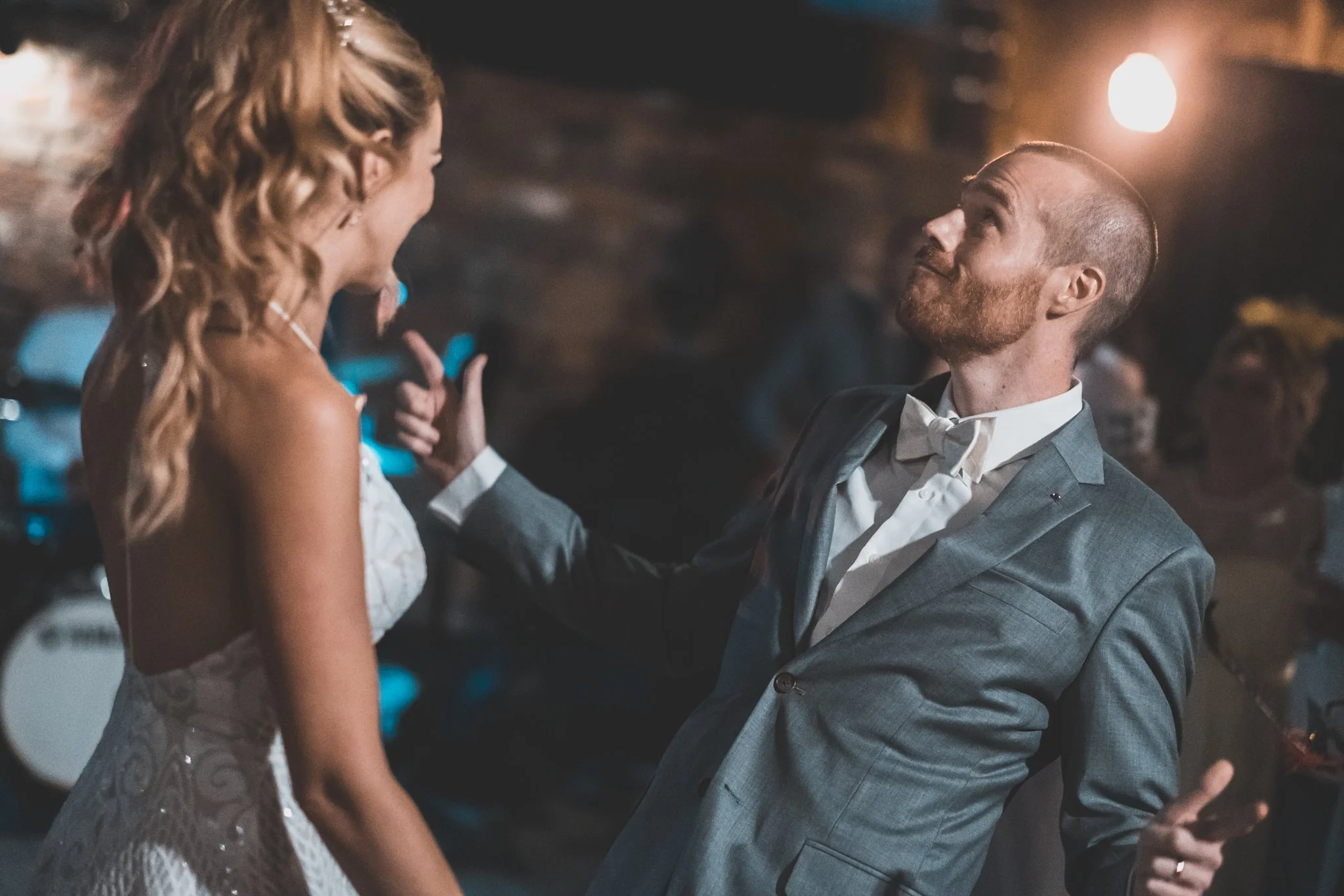 A man in a gray suit and white bow tie sitting down and looking up at a woman with long curly hair in an elegant white dress, at a dimly lit event or party.