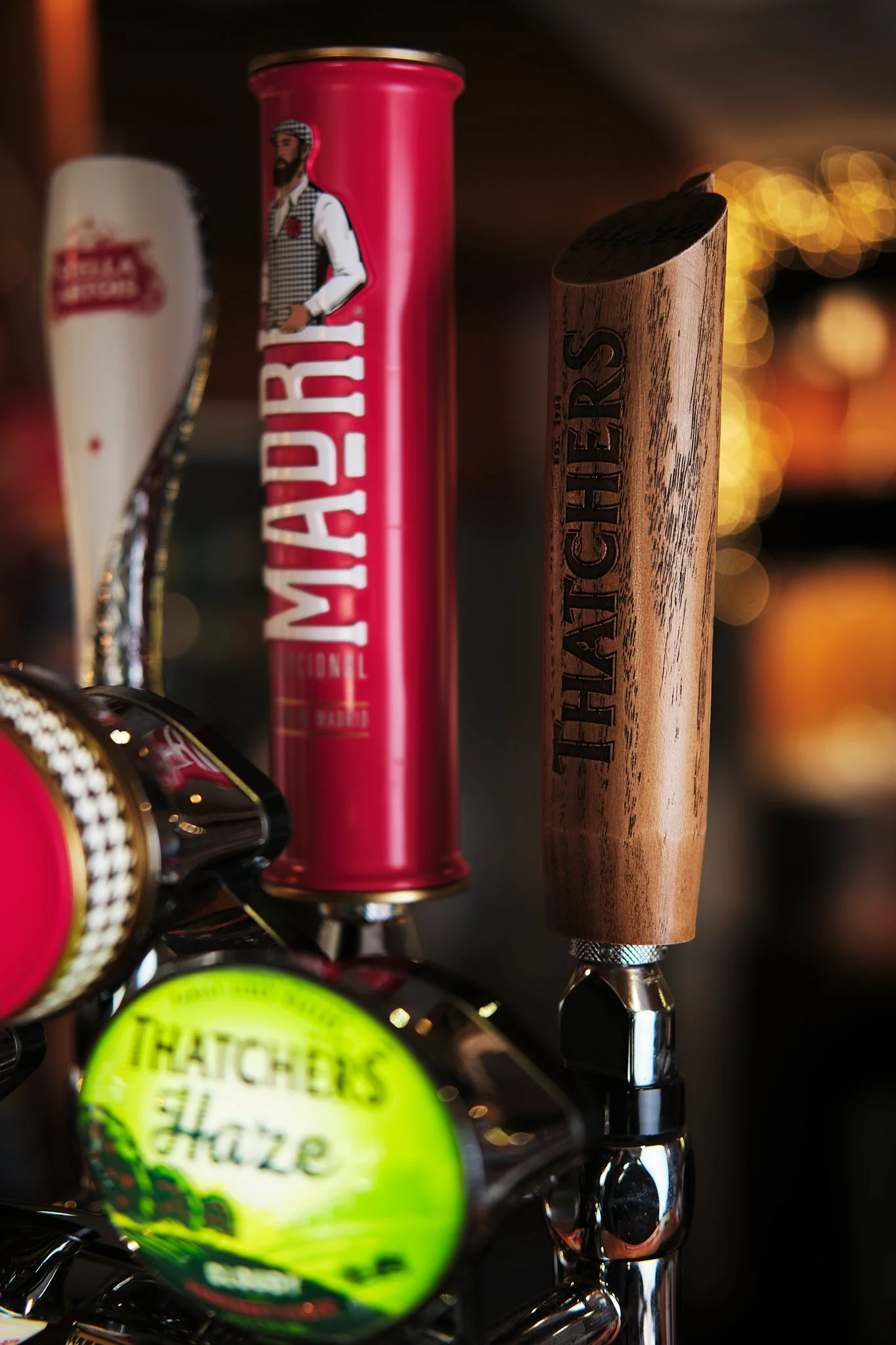 Close-up of beer tap handles on a bar, including a red handle labeled "Madira" with a bearded man illustration, a wooden handle labeled "Hatchers," and a partially visible handle with a round green label that reads "Thatchers haze."