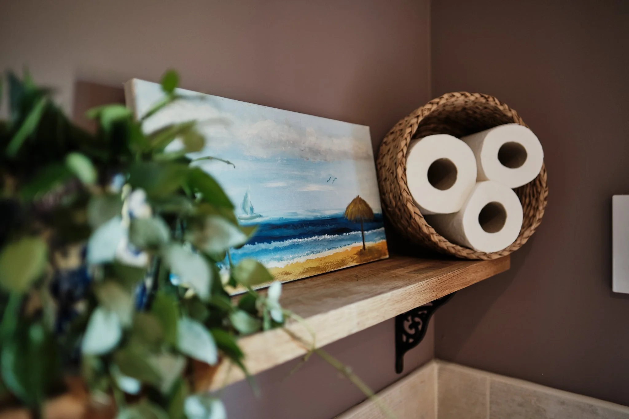 A shelf with a potted green plant, a seascape painting of a beach with umbrellas, and a woven basket holding three rolls of toilet paper.