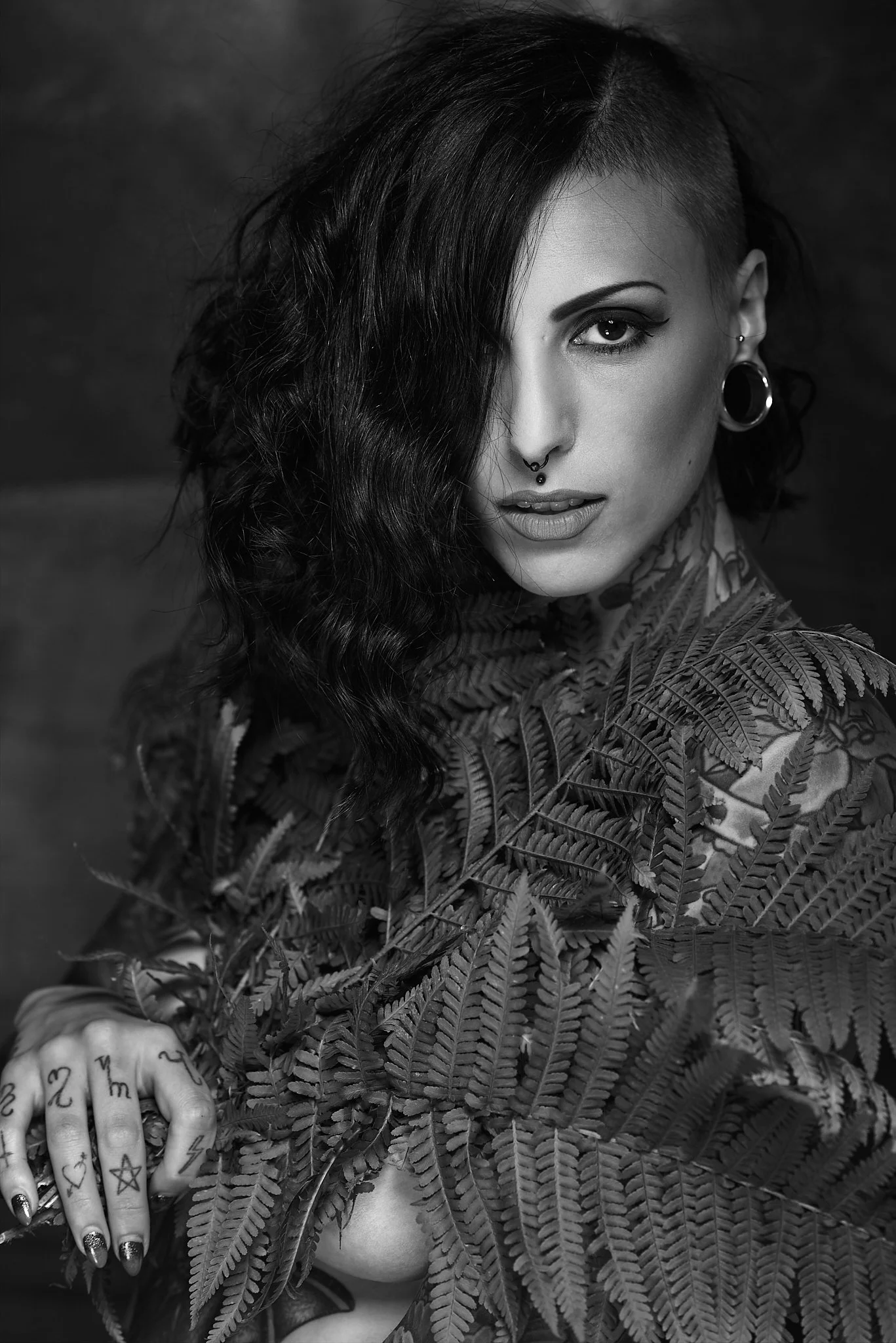 A black and white portrait of a woman with dark, wavy hair covering one eye, multiple ear piercings, and tattoos on her fingers. She is holding fern leaves close to her chest, with a partially visible shoulder and bare skin, and a textured background