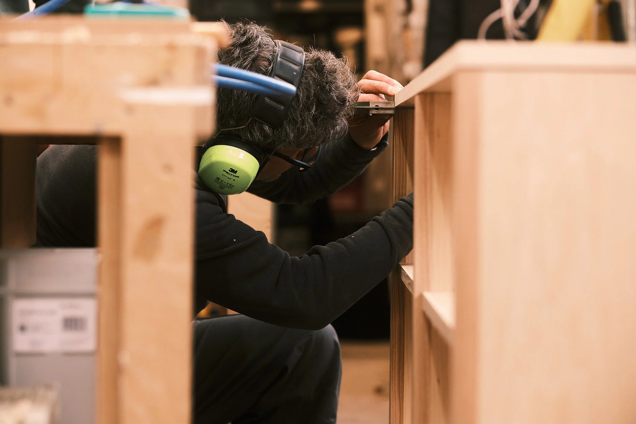 A person wearing green ear protection and glasses, working on a wooden piece in a workshop.