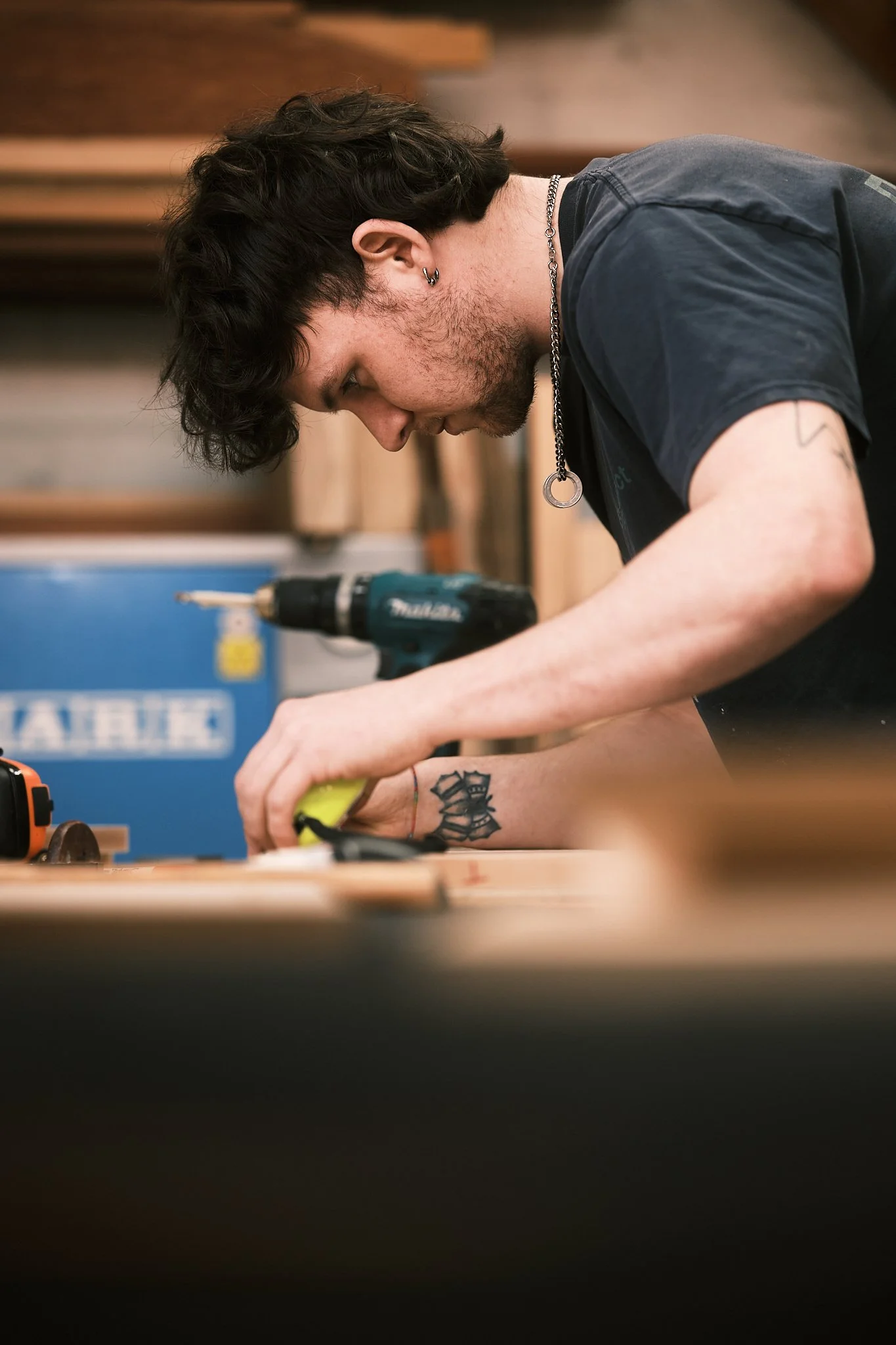 A young man with dark hair and tattoos is working with a power drill in a woodworking shop.