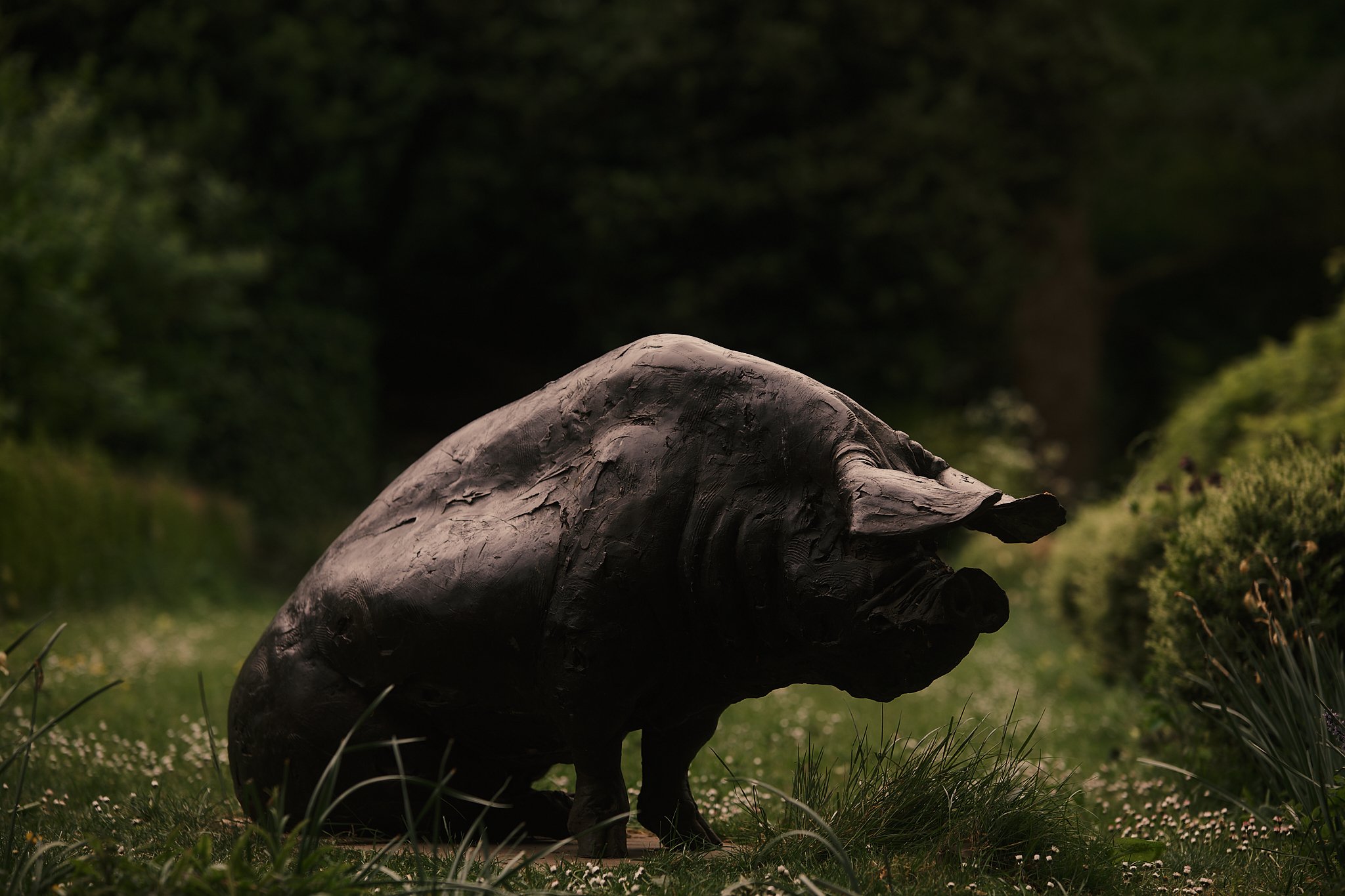 A dark, artistic sculpture of a creature resembling a hybrid between a dog and a pig, placed outdoors on grass with trees and greenery in the background. Hotel interior photography at THE PIG-in the Cotswolds