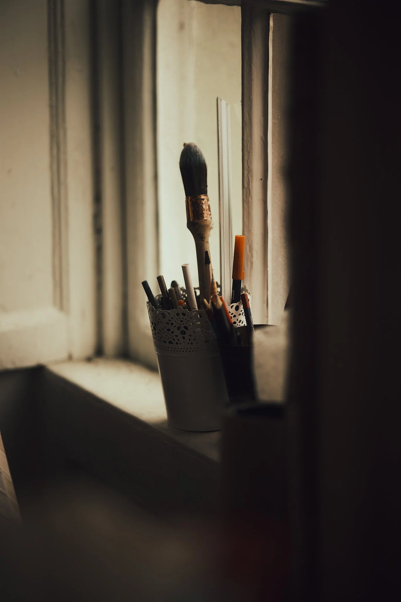 Paintbrushes and pens in a white perforated container on a windowsill, with sunlight illuminating the scene.