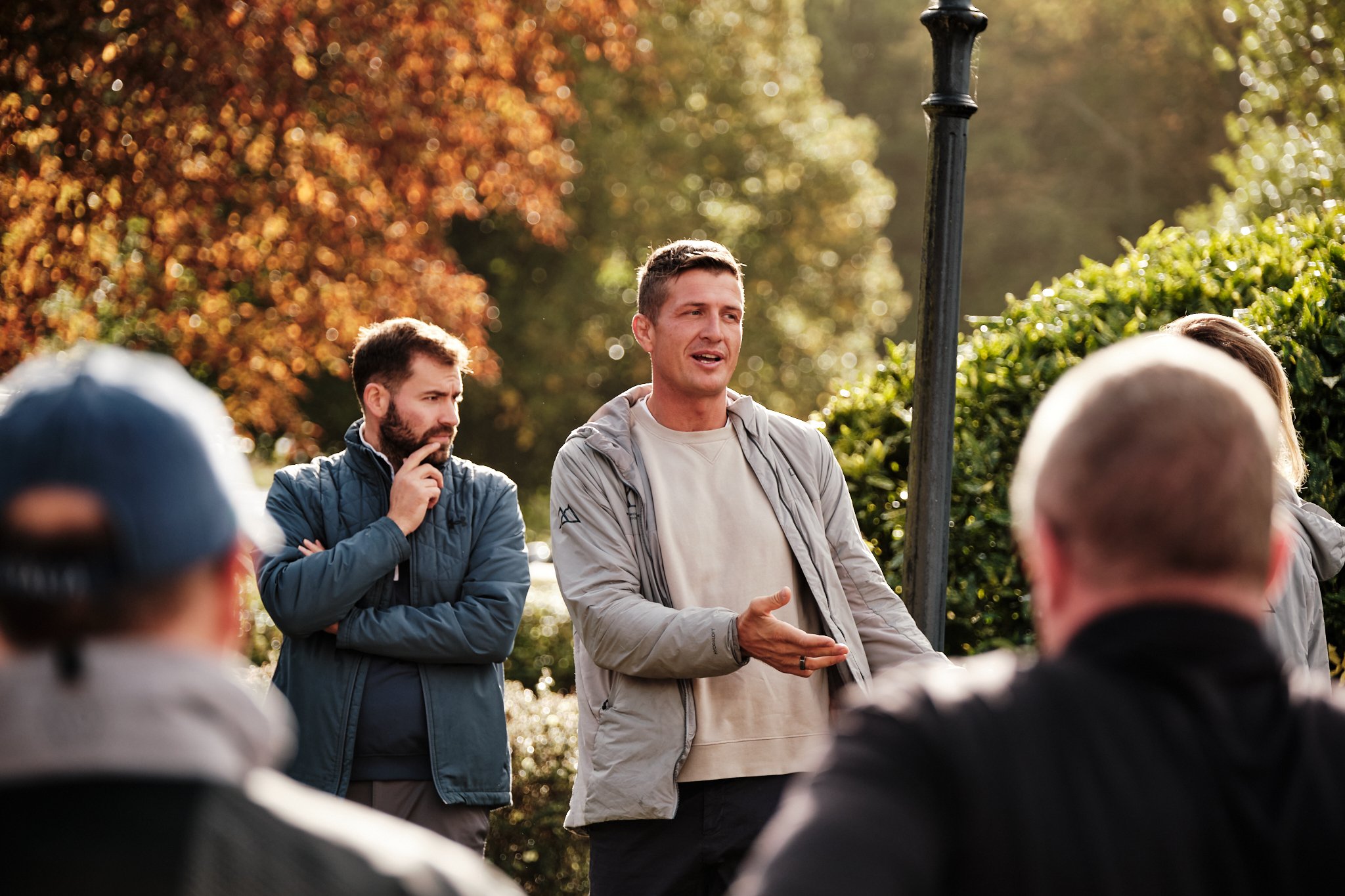 A man is speaking to a group of people outdoors during autumn, with trees and bushes with orange and green leaves in the background. Others are listening, some with thoughtful expressions.