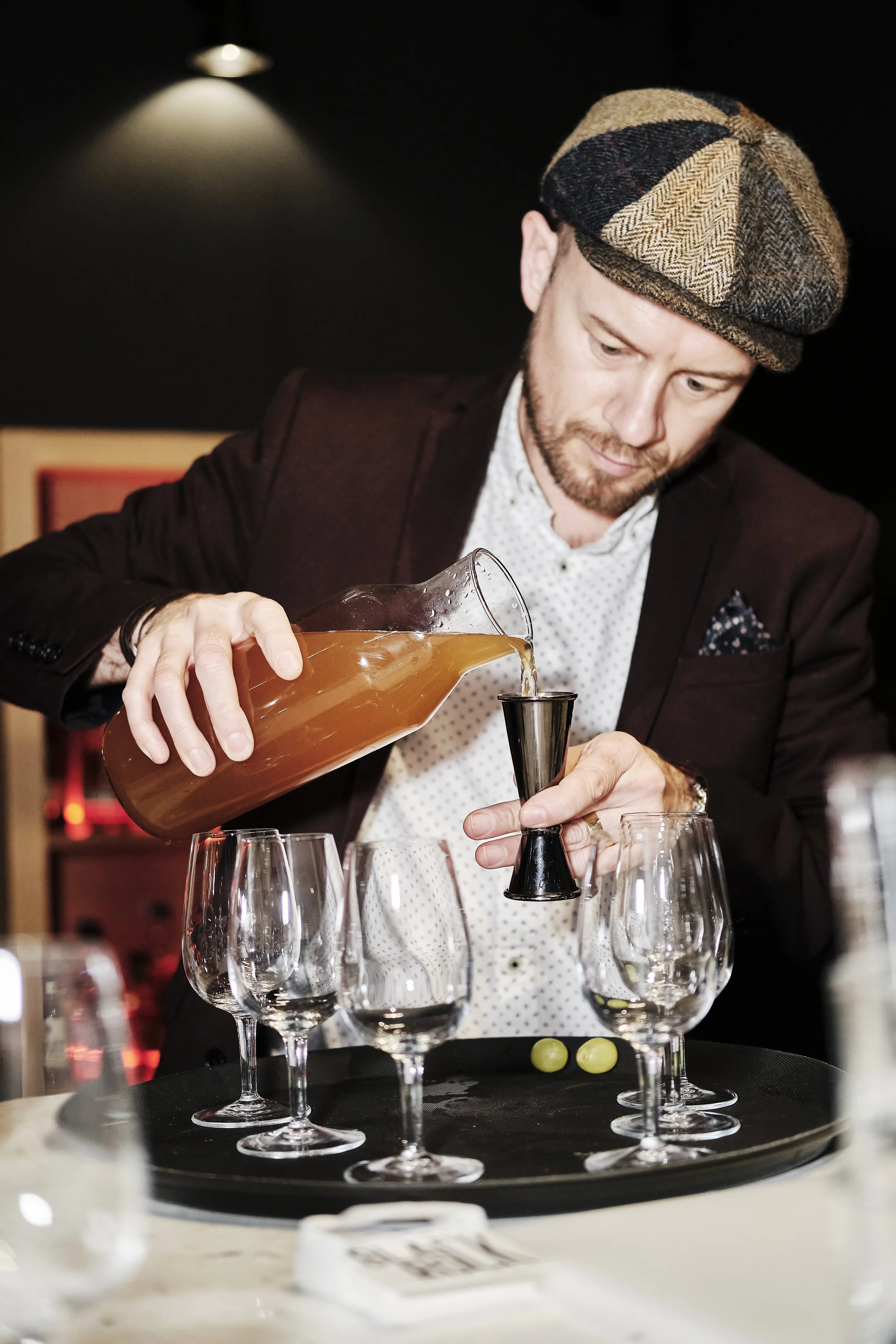 A man in a tartan flat cap and dark blazer pouring a liquid from a glass decanter into a jigger, with several empty wine glasses and green grapes on a round black tray on a table.