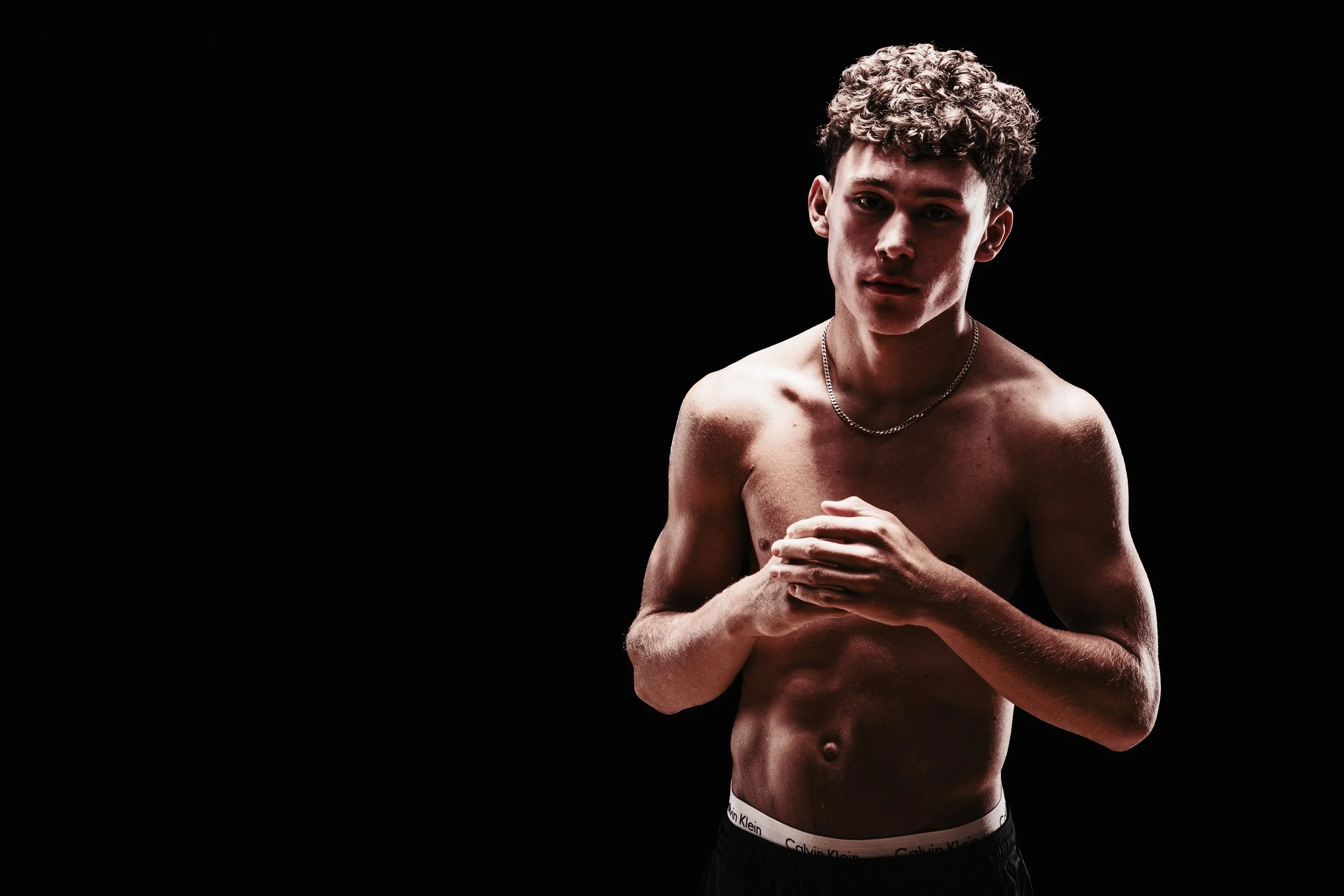 A shirtless young man with curly hair and a necklace stands against a dark background, looking at the camera.