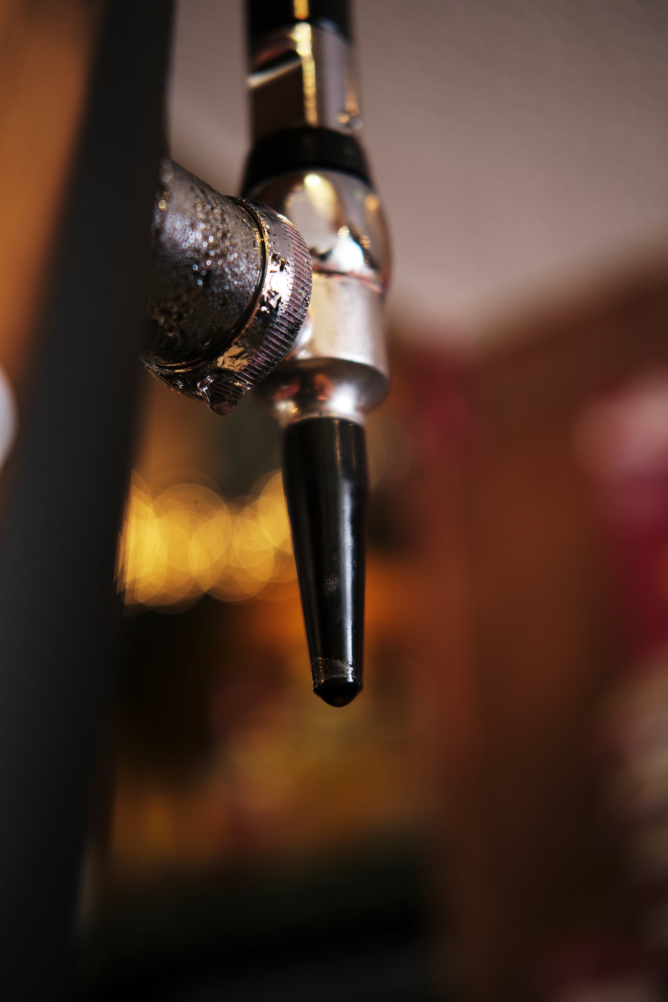 Close-up of a beer tap with condensation, attached to a shiny metal faucet, against a blurred background with warm lights.