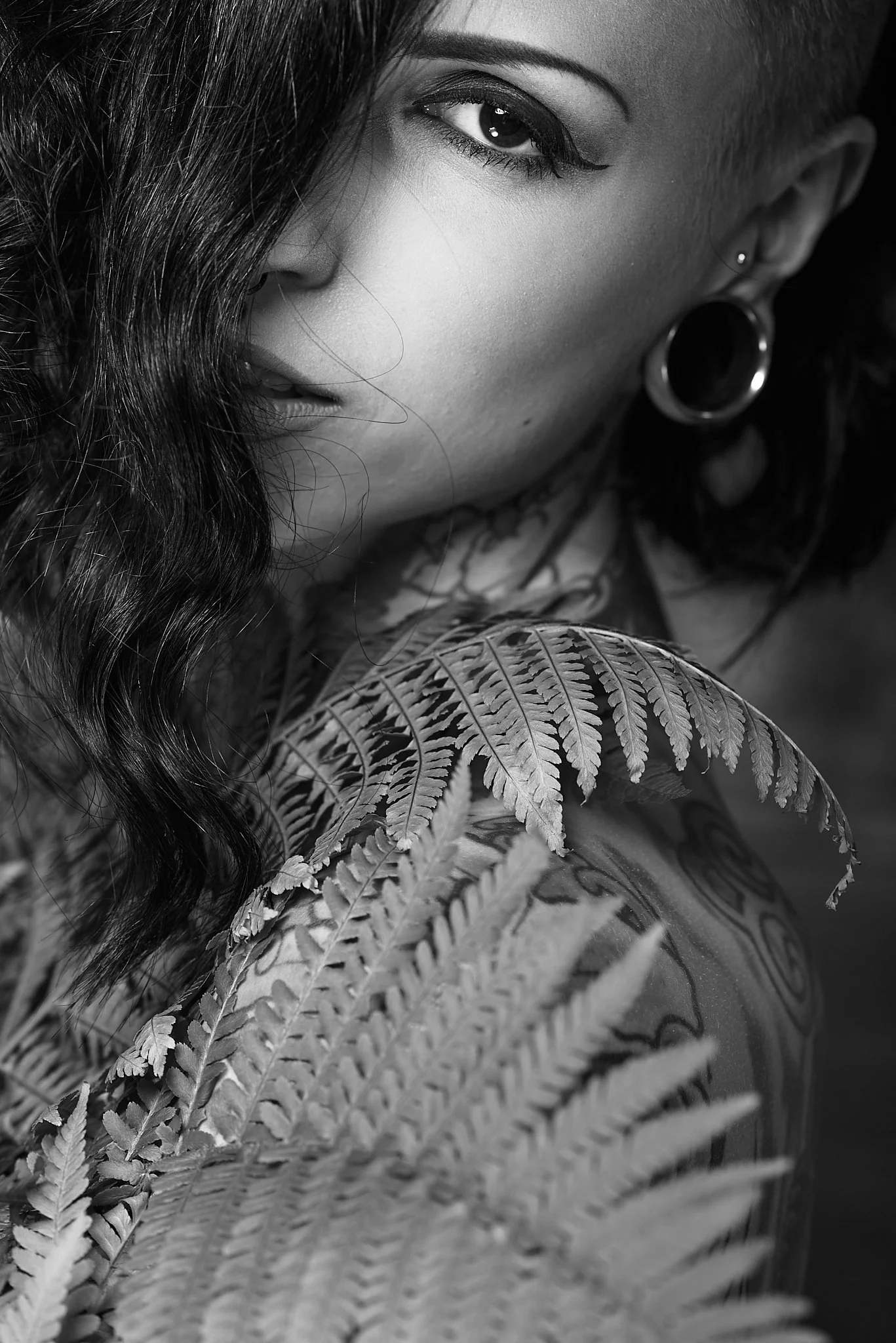 A woman with dark curly hair, piercings, and tattoos, partially obscured face, holding a fern plant with detailed leaves.